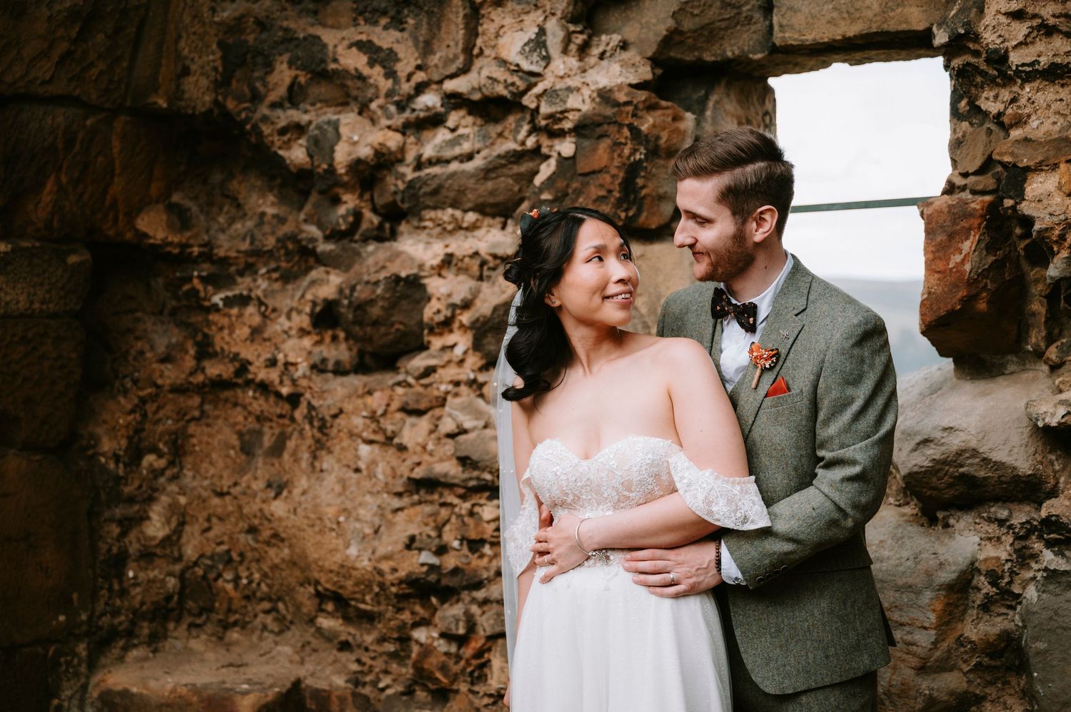 A romantic wedding couple embraces against a rustic stone wall with natural lighting streaming through an opening.