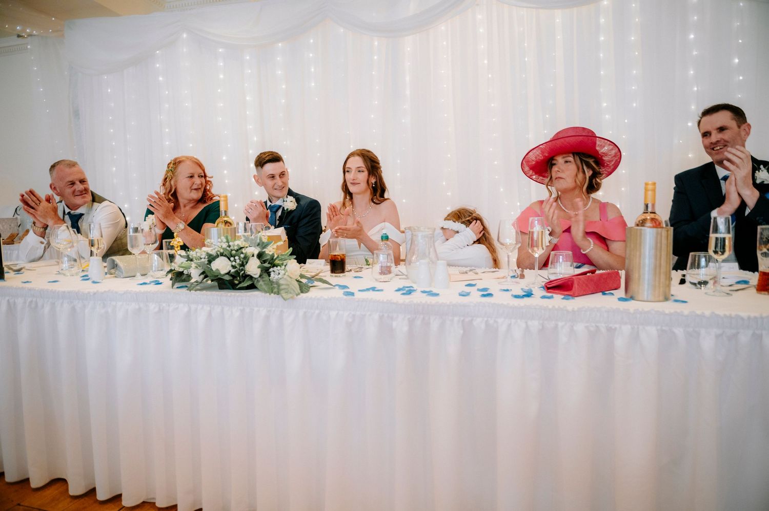 Long head table at wedding reception with guests seated against white backdrop with string lights.