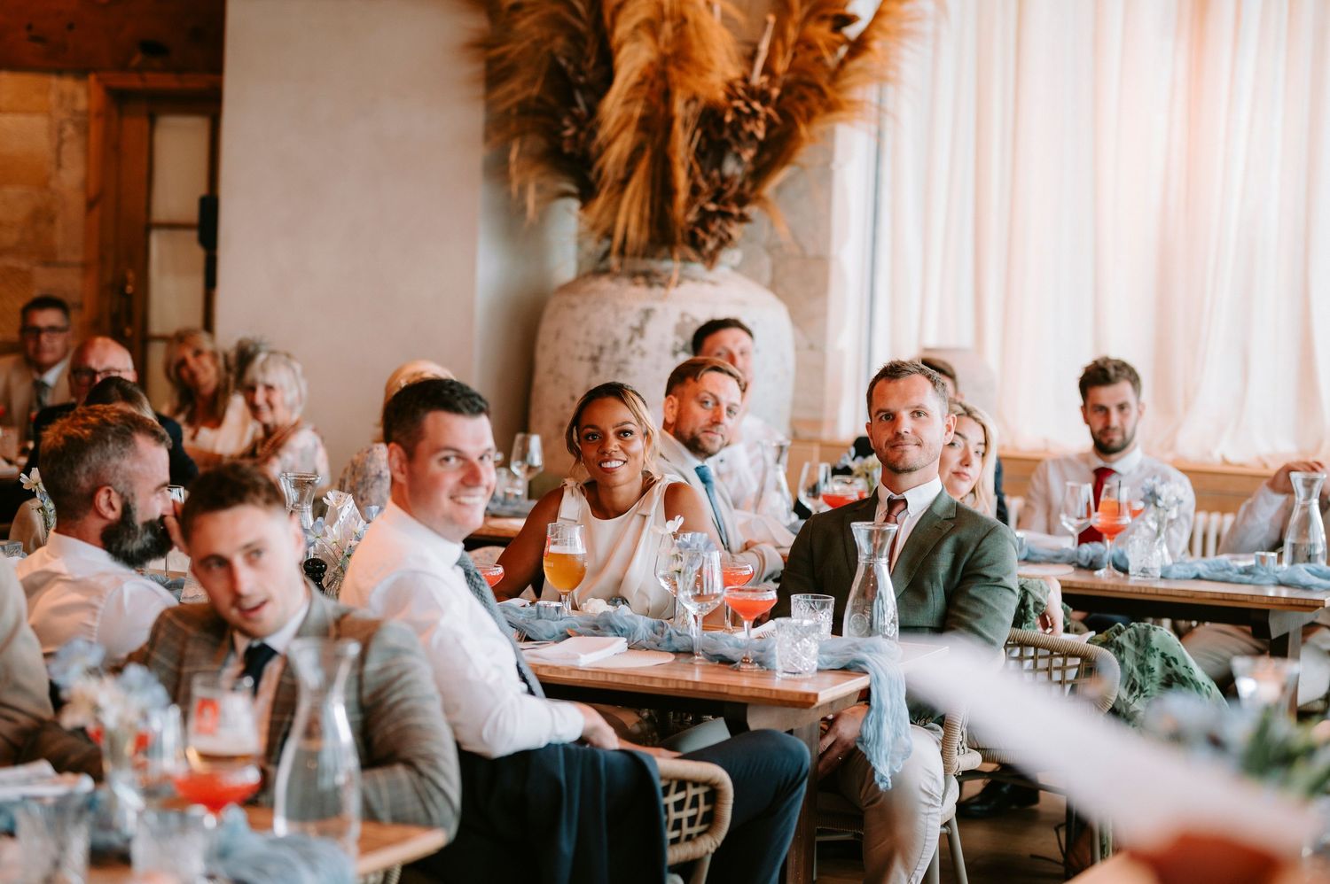 Wedding guests seated at long tables in a bright reception venue react with laughter and smiles during speeches.