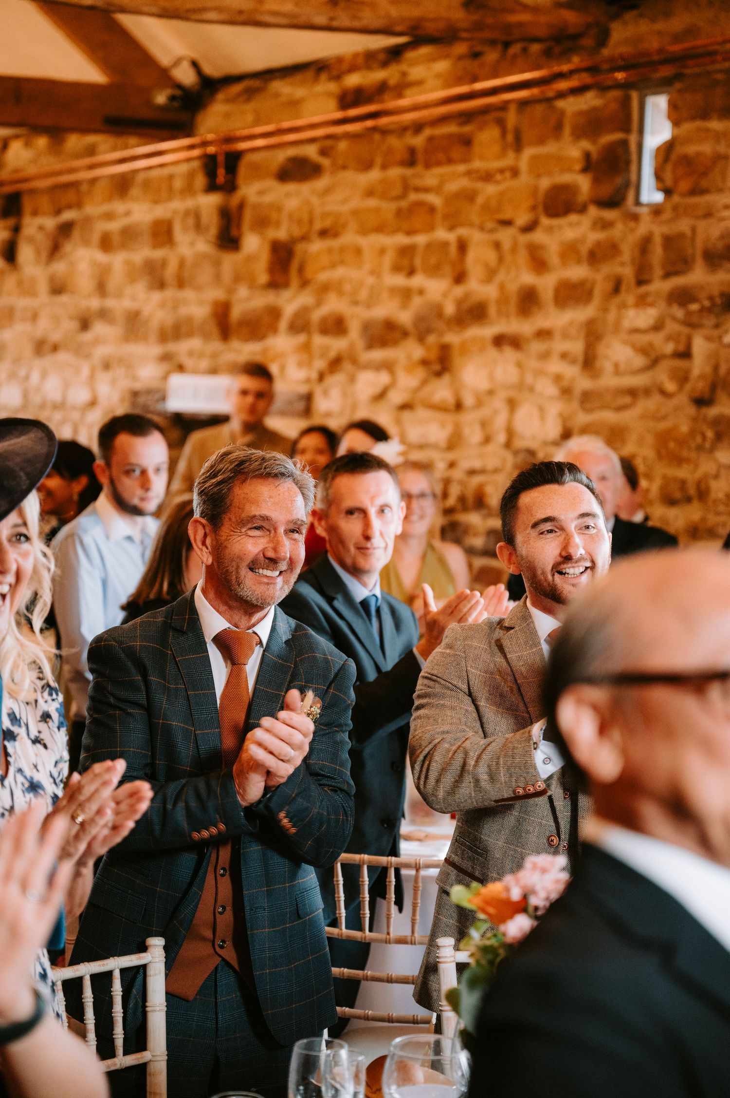 Wedding guests laugh and smile during a reception in a rustic brick-walled venue.