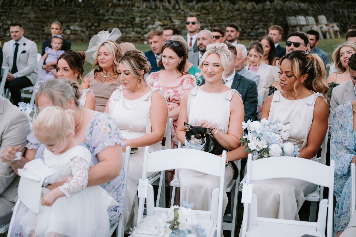 Wedding guests seated in white chairs outdoors react emotionally during ceremony.