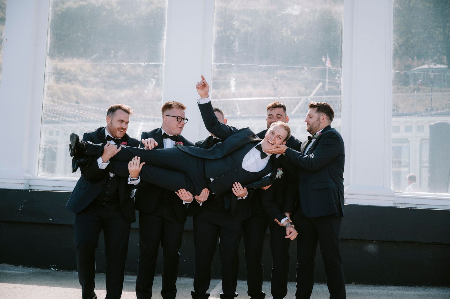 Groomsmen in black tuxedos posing casually against a bright architectural backdrop.