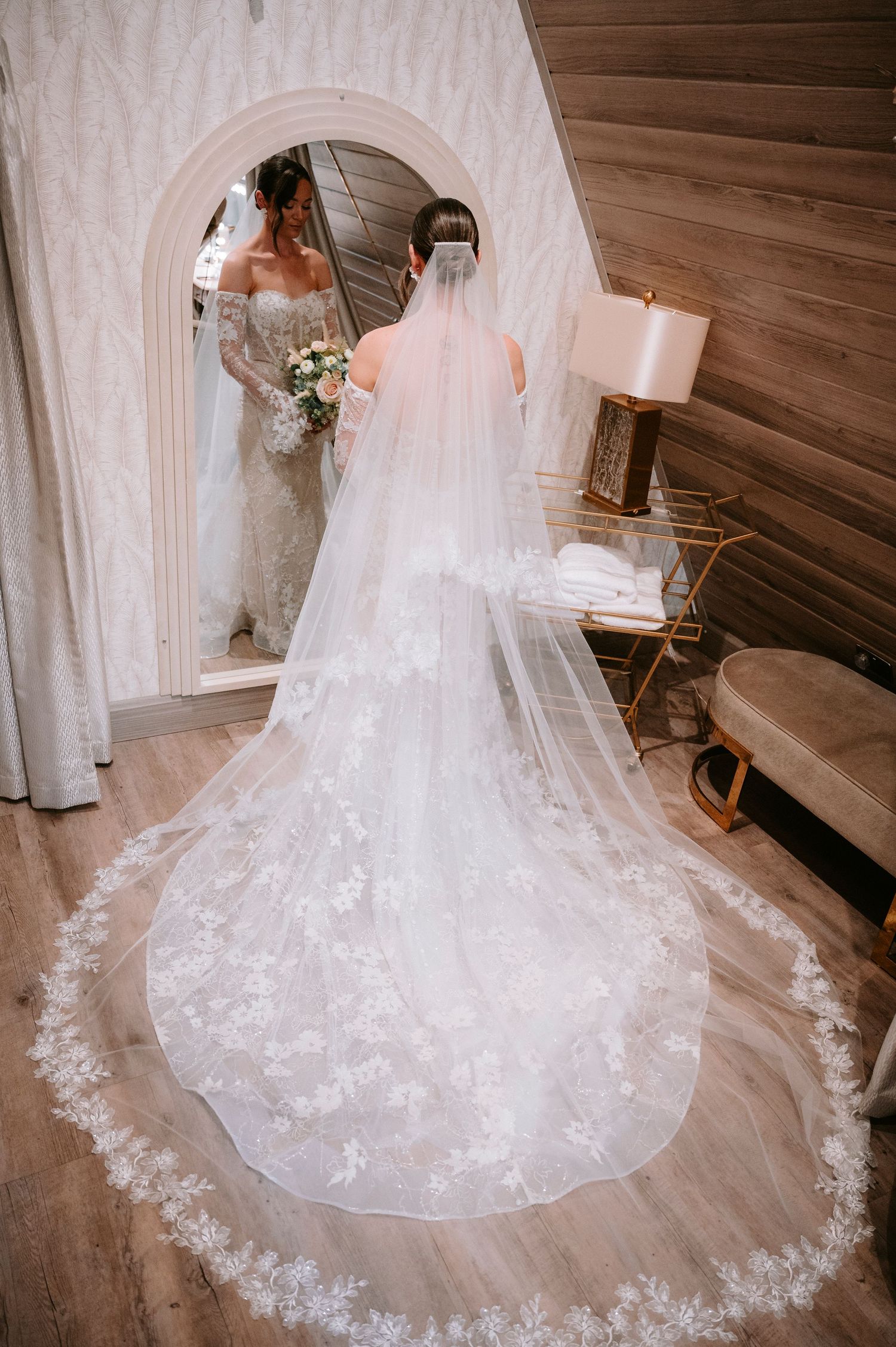 A stunning bridal reflection in an arched mirror showcases an intricate lace veil trailing on wooden floors.