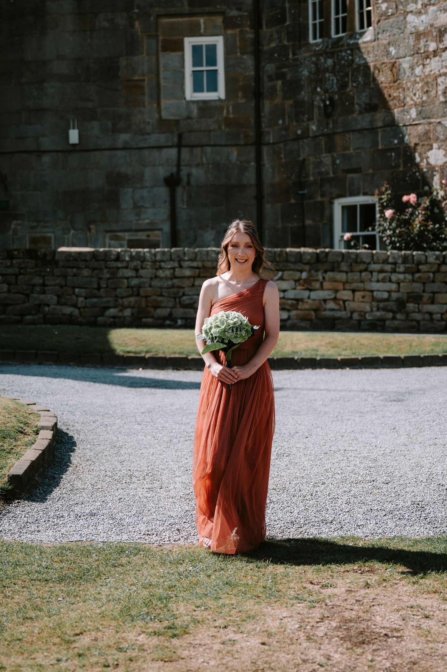 A bridesmaid in a coral-colored dress carries a bouquet while walking on gravel outside a stone building.