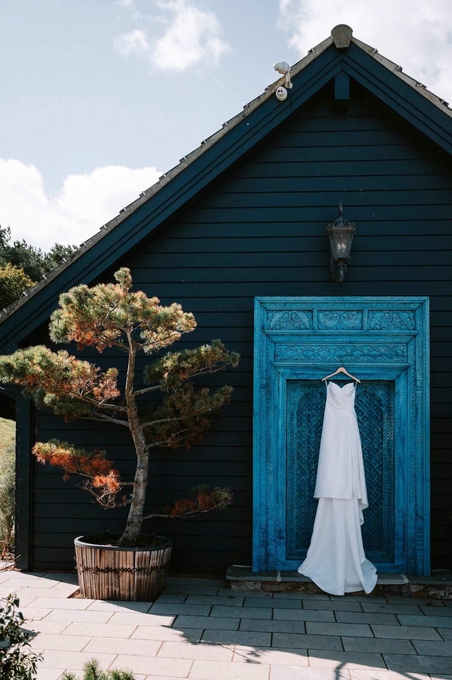 A white wedding dress hangs elegantly against a dark blue wooden building with ornate turquoise doors.