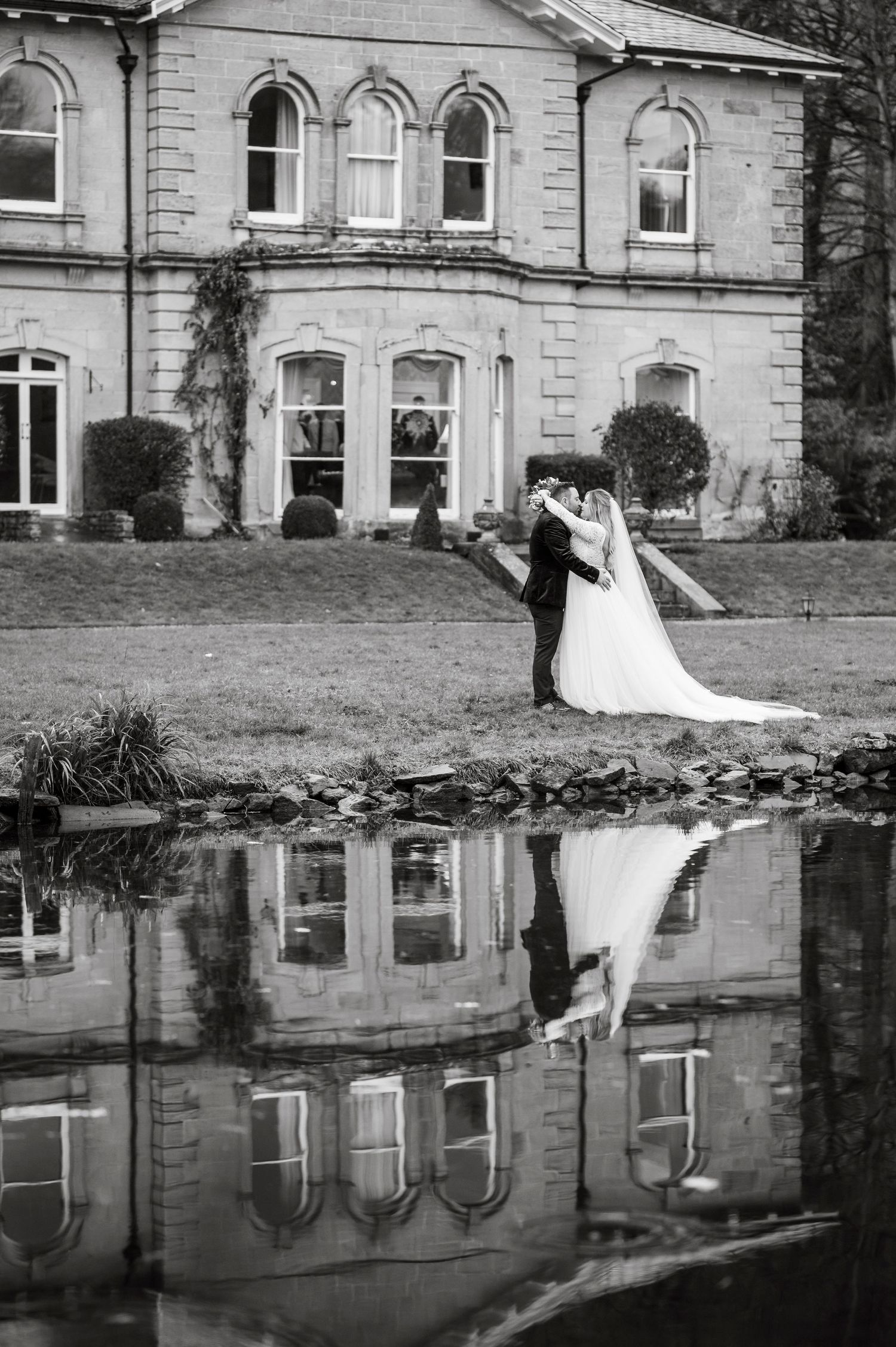 Black and white photo of newlyweds reflected in a pond beside a stone mansion.