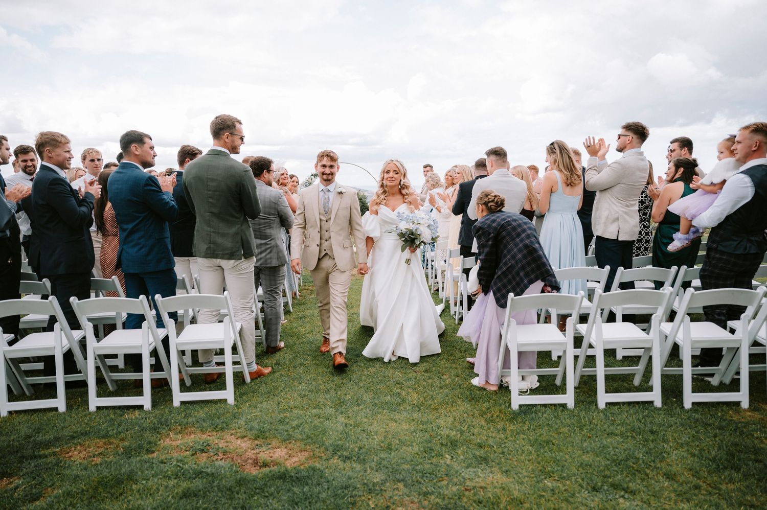 Outdoor wedding ceremony with rows of white chairs set up on a grassy hill under cloudy skies.
