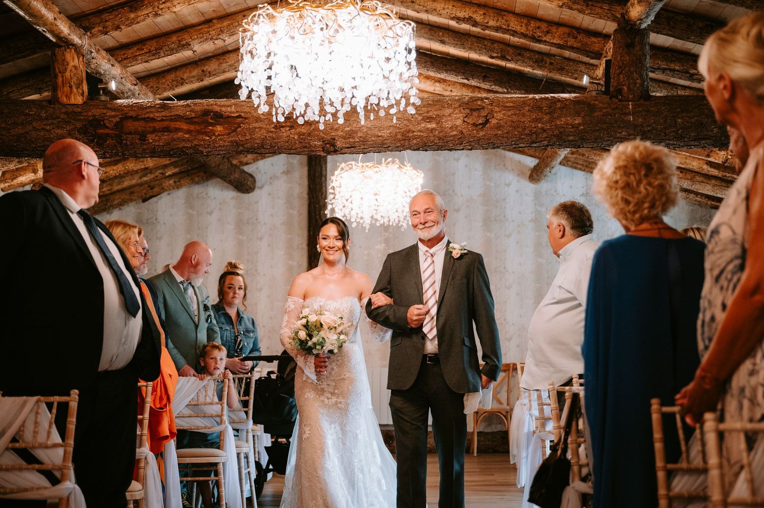 Wedding ceremony in progress inside a rustic wooden venue with crystal chandelier overhead.
