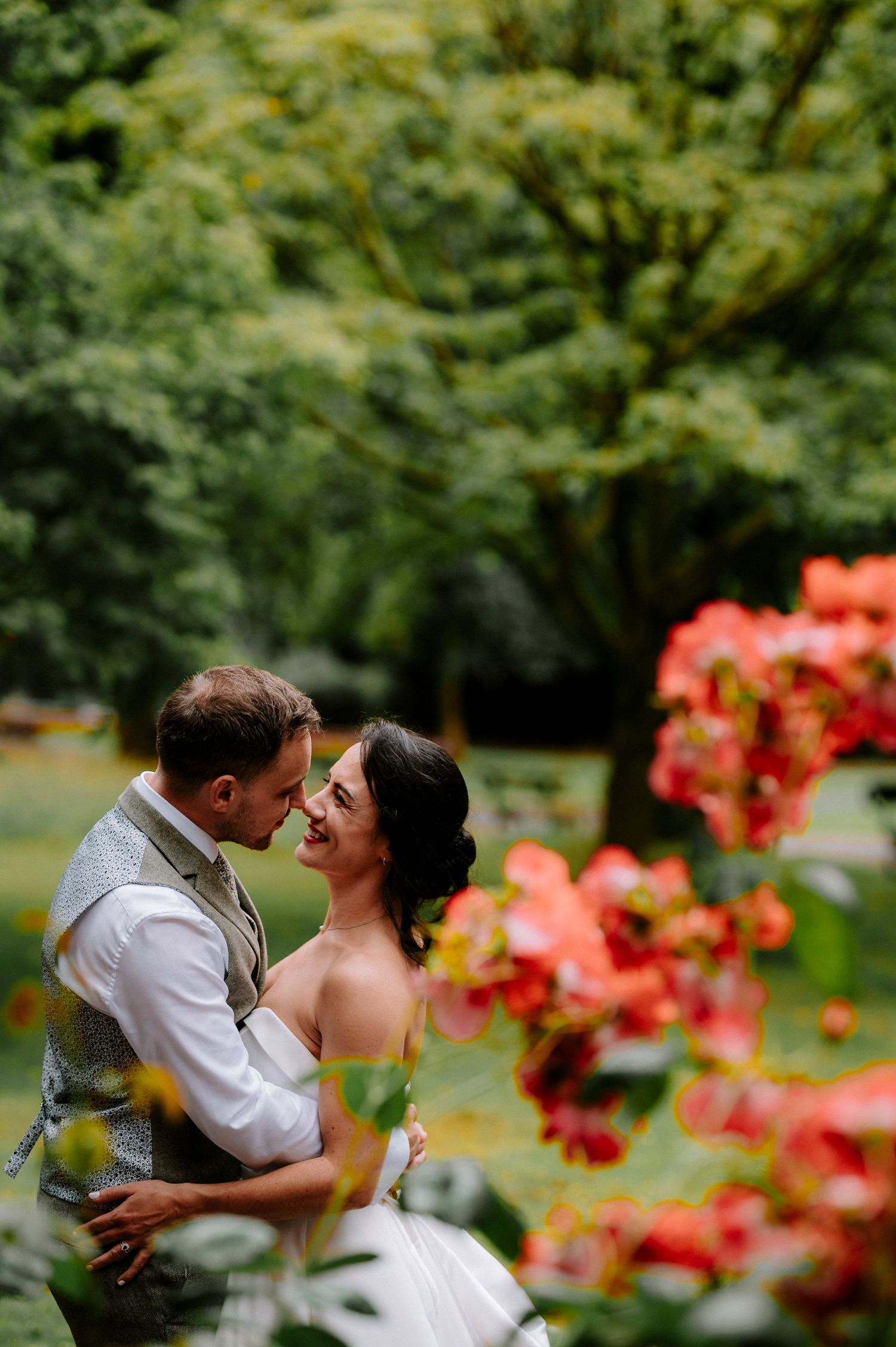 A tender wedding moment captured in a garden with vibrant coral flowers in the foreground.