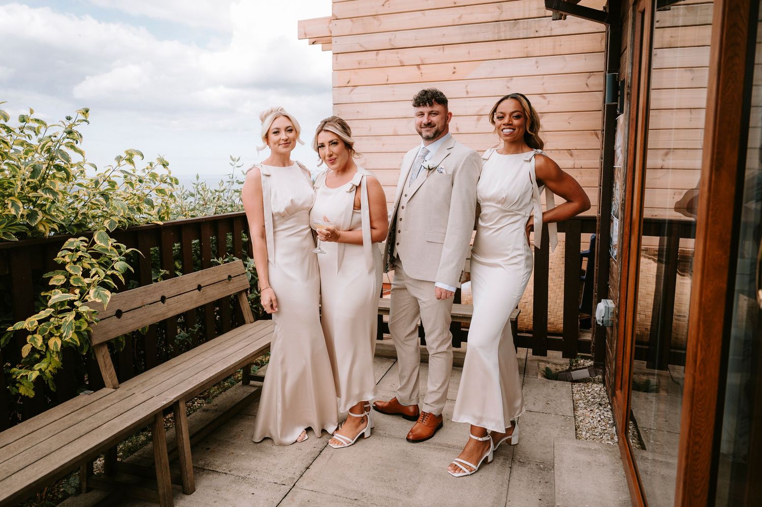 Wedding party in white and beige attire poses together on an outdoor deck with greenery in background.