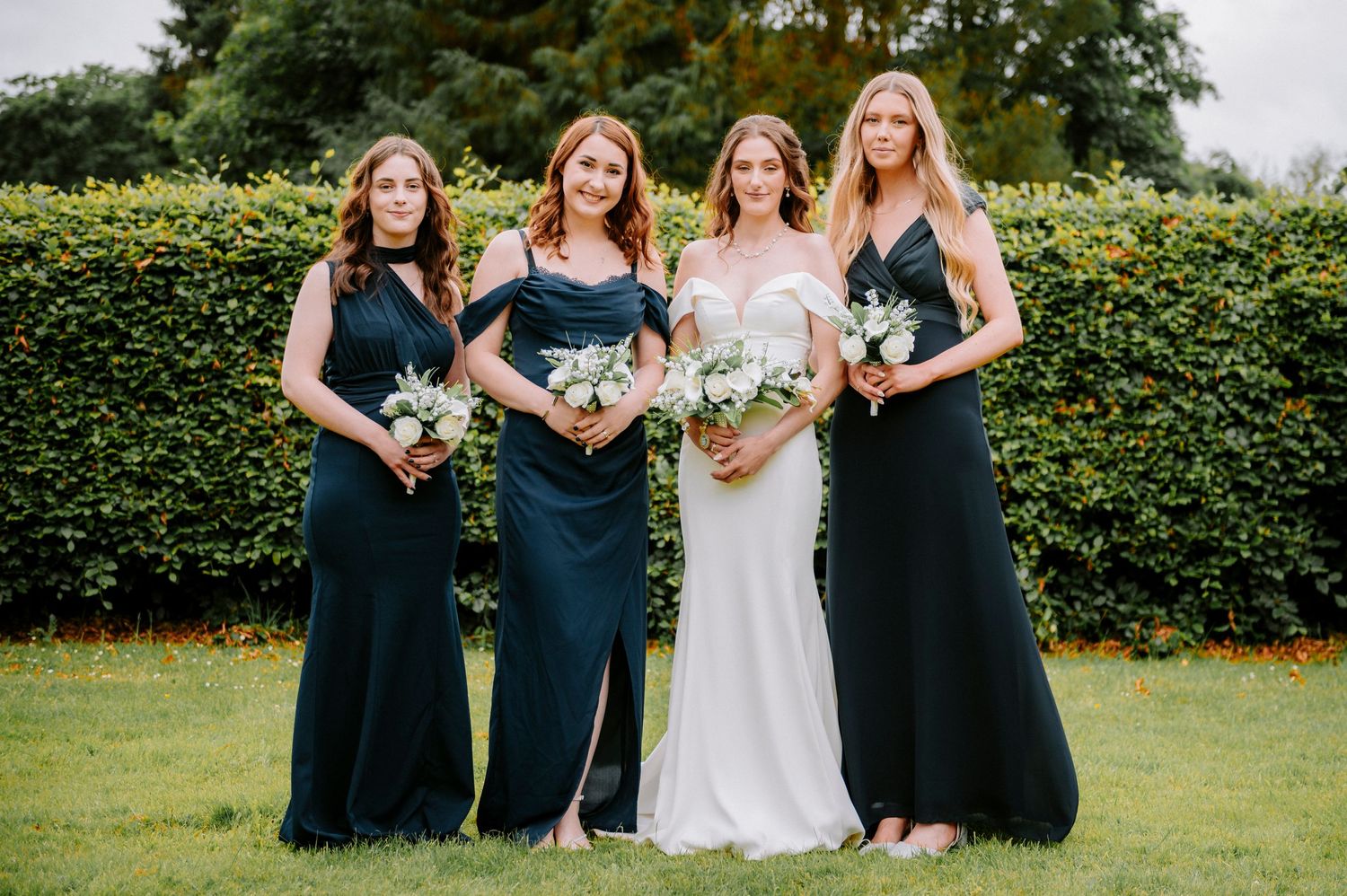 Bridal party in navy dresses poses outdoors against a backdrop of hedges.