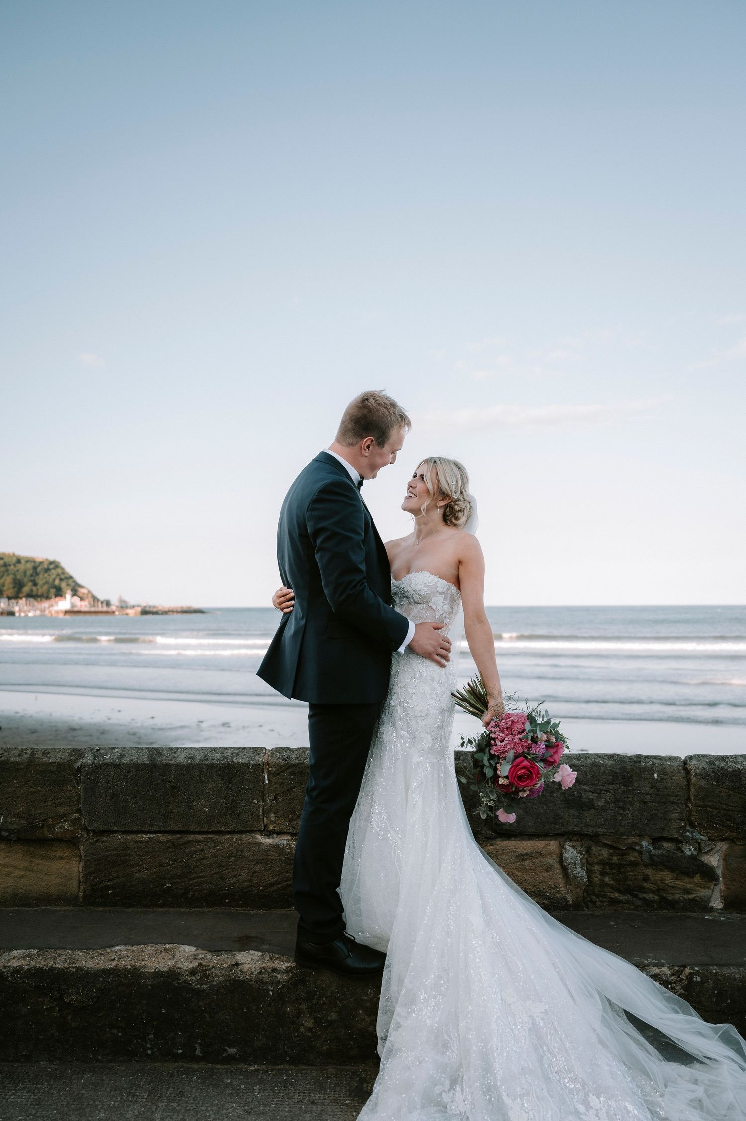 Seaside wedding portraits with white dress against dramatic ocean and stone wall backdrop.