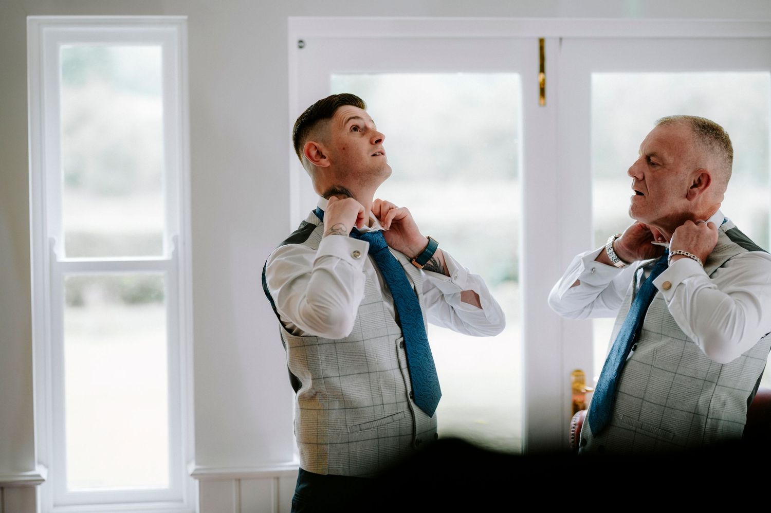 Two men in matching waistcoats adjusting their ties while getting ready.