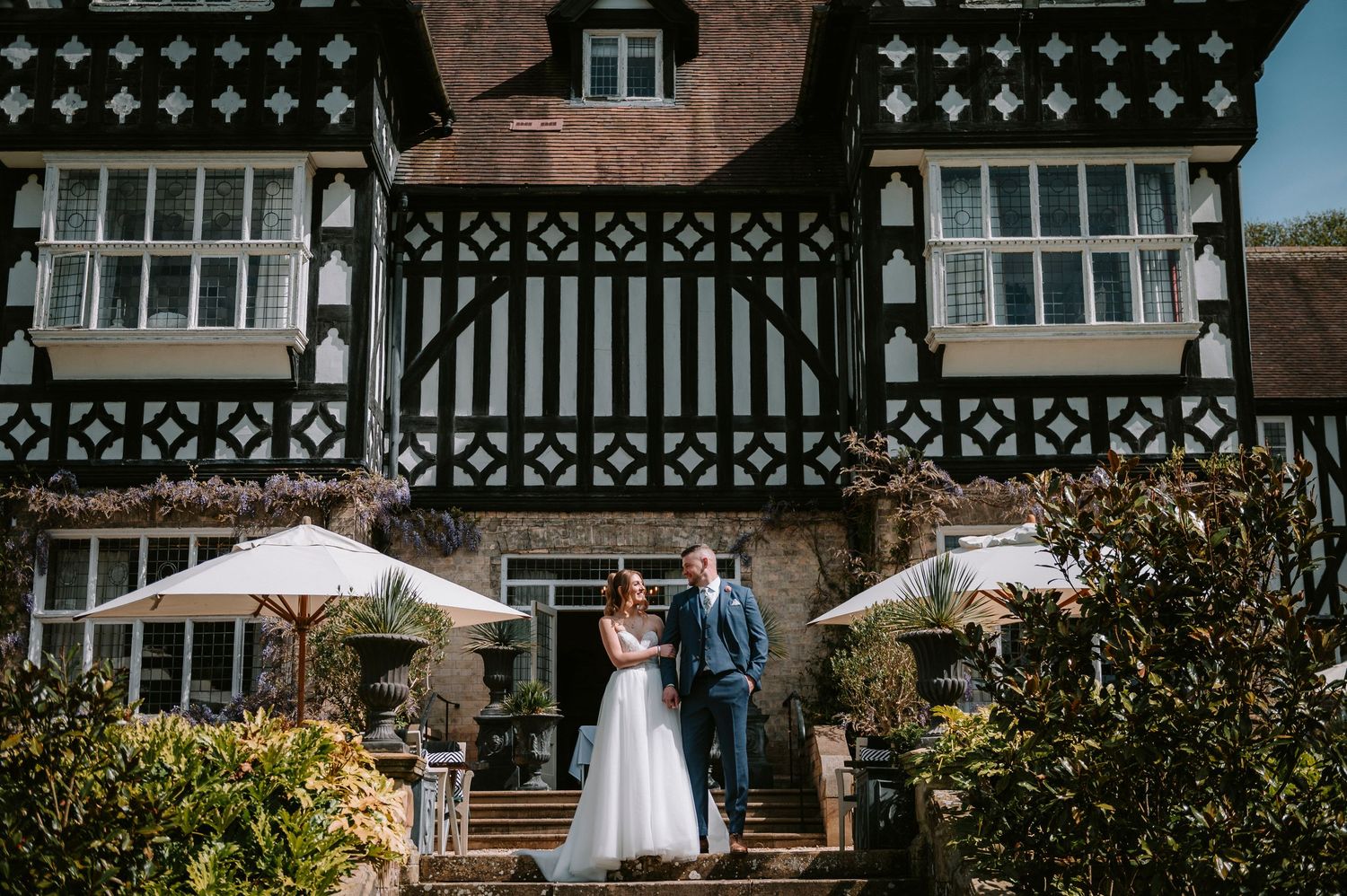 A wedding couple poses before a Tudor-style building with distinctive black and white architectural details.