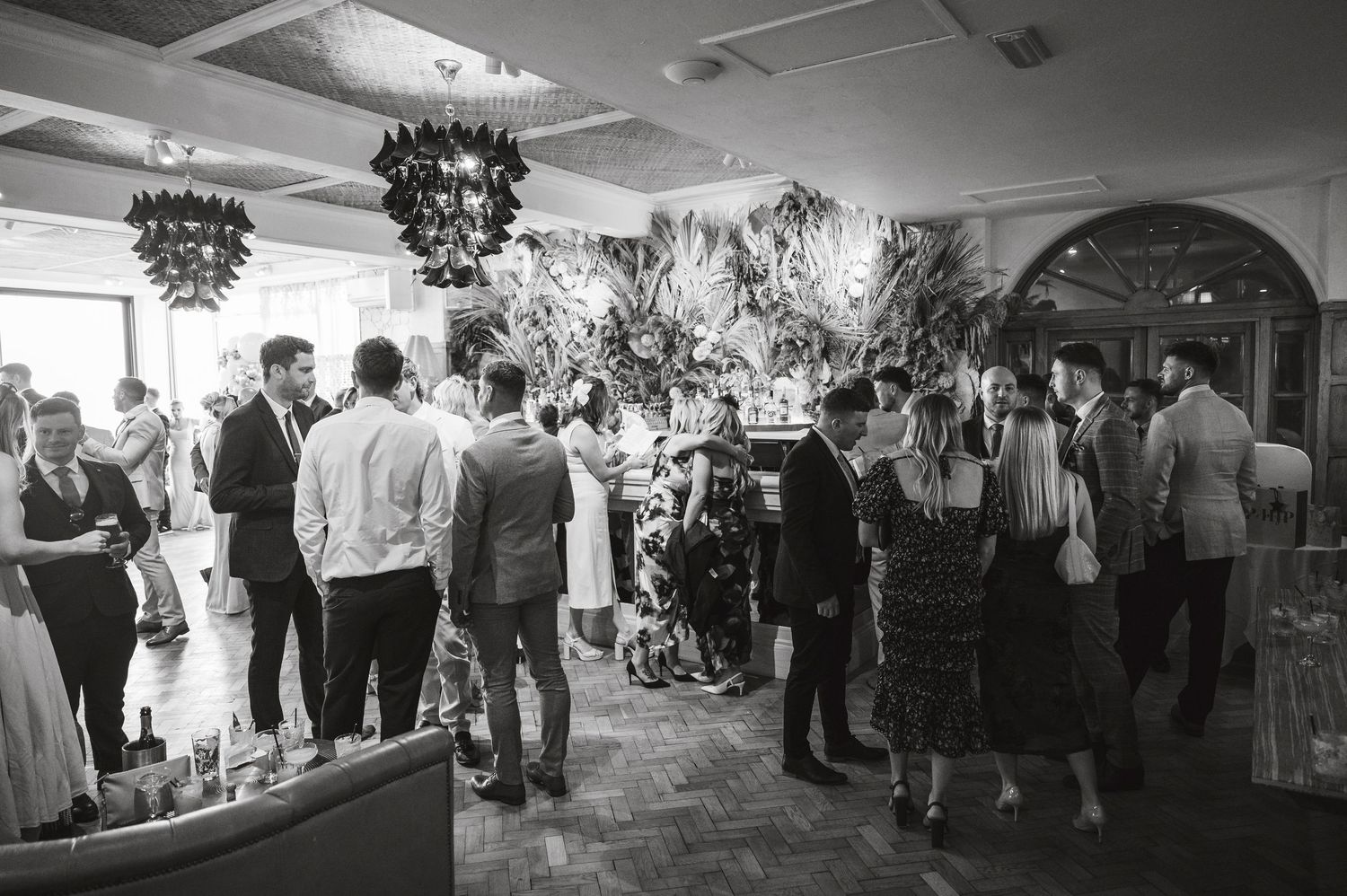 Black and white panoramic shot of wedding reception with guests mingling in an indoor venue.