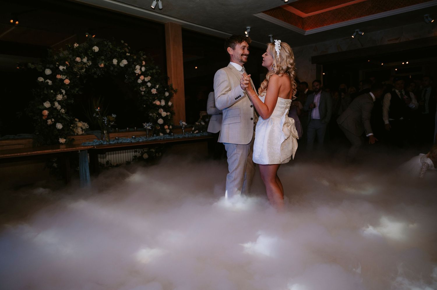 Couple dances on misty dance floor with floral arch in background at evening reception.