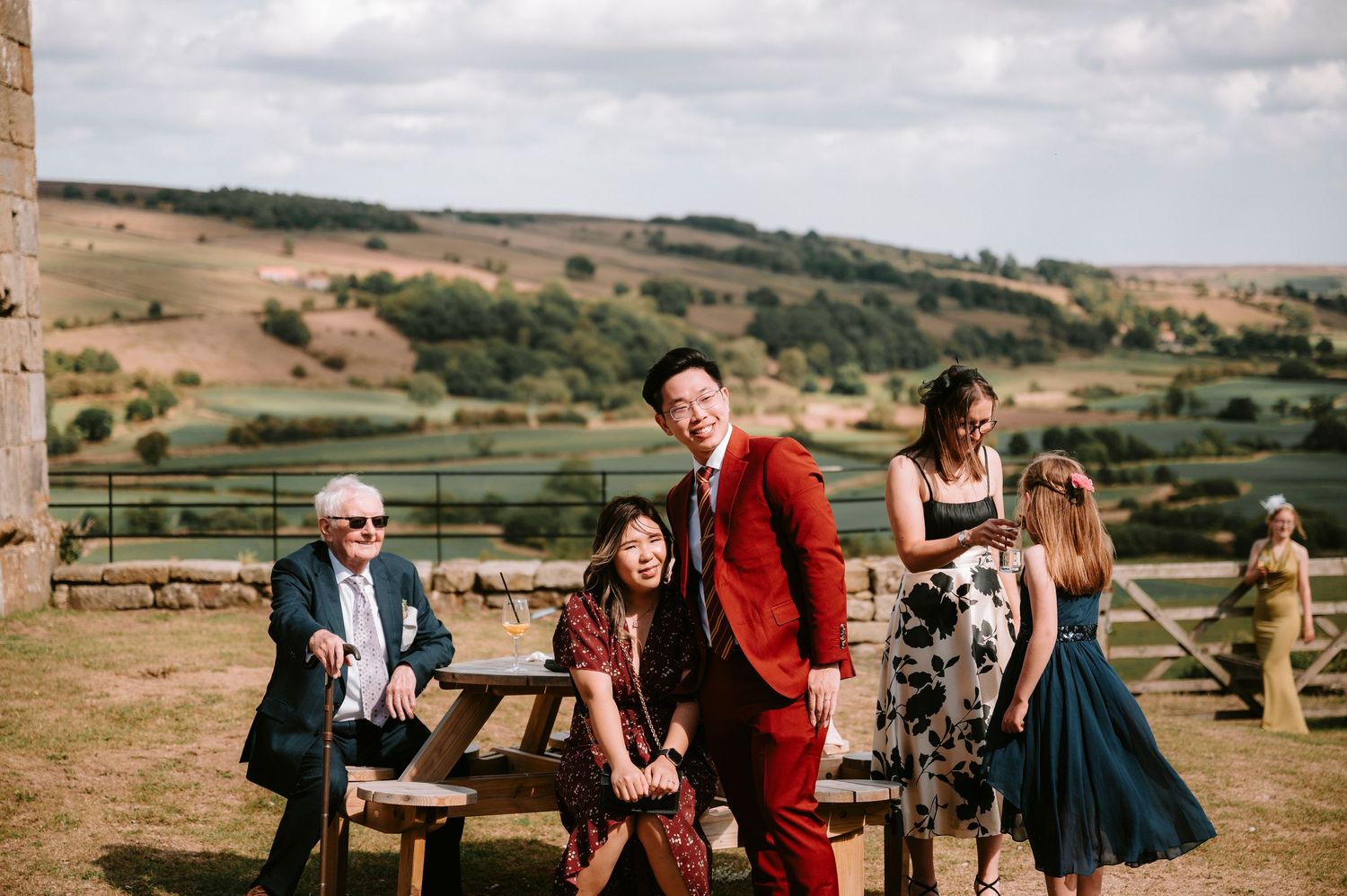 Wedding guests in formal attire enjoying themselves at an outdoor reception with rolling hills in background.