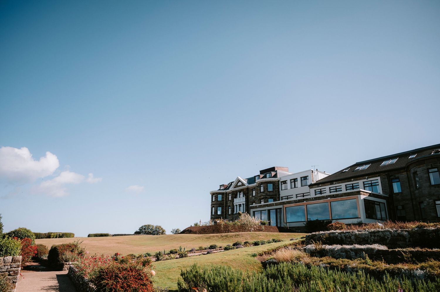 Modern coastal house with large windows perched on a hilltop overlooking natural landscape against blue sky.