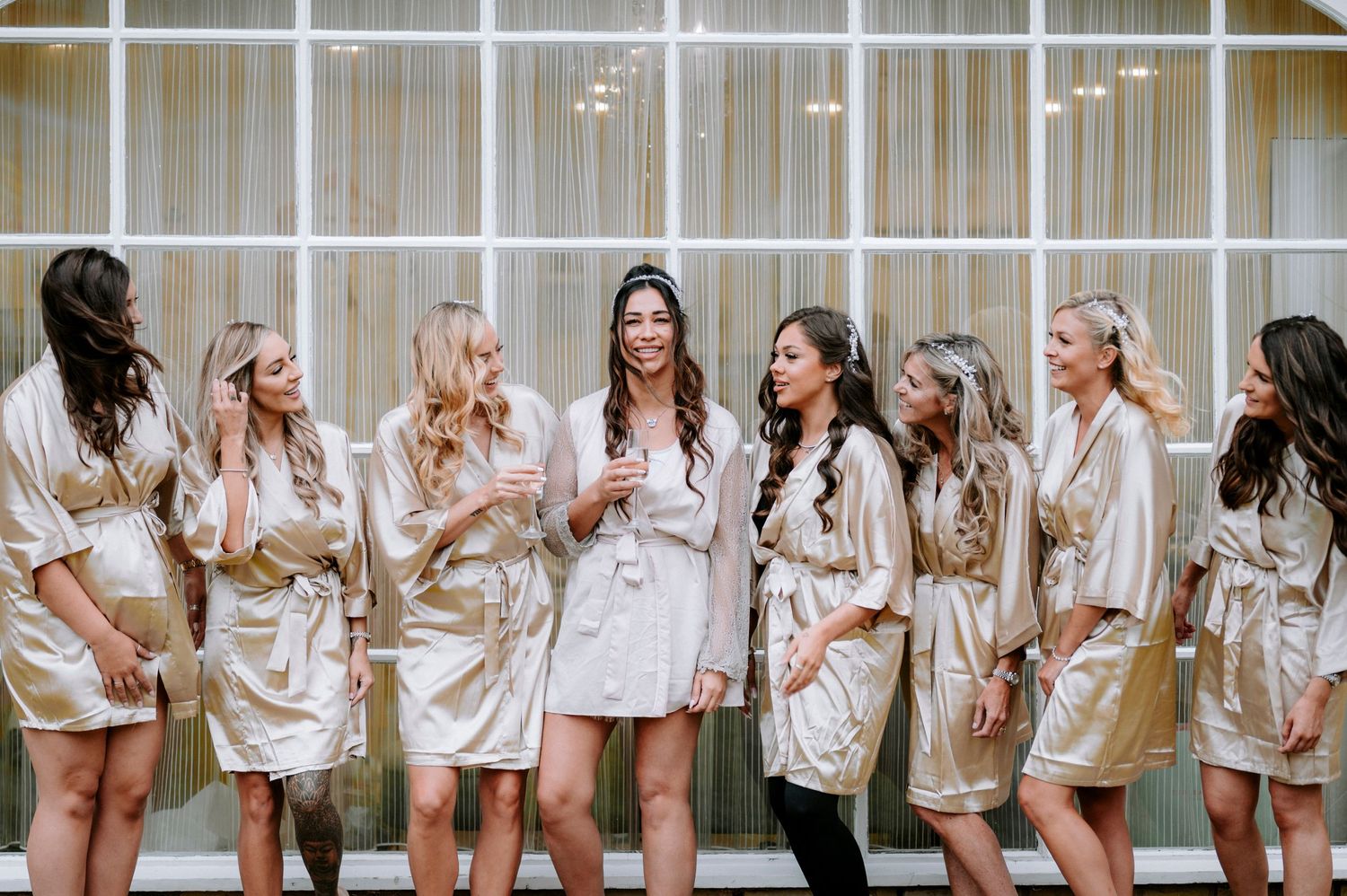 Row of bridesmaids wearing matching white satin robes lined up against a window.
