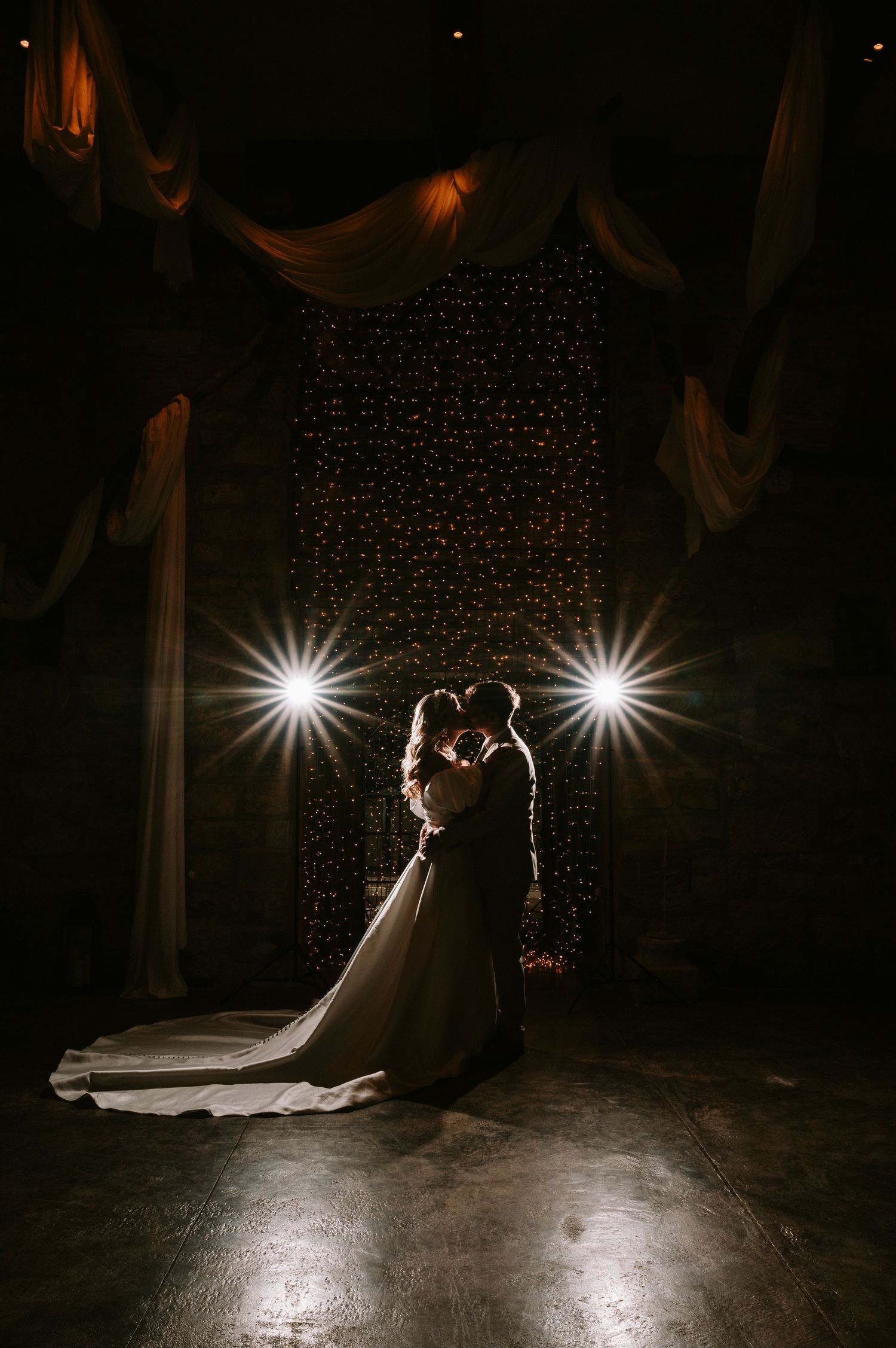 Silhouetted couple shares romantic dance under twinkling fairy lights and spotlights in dark venue.