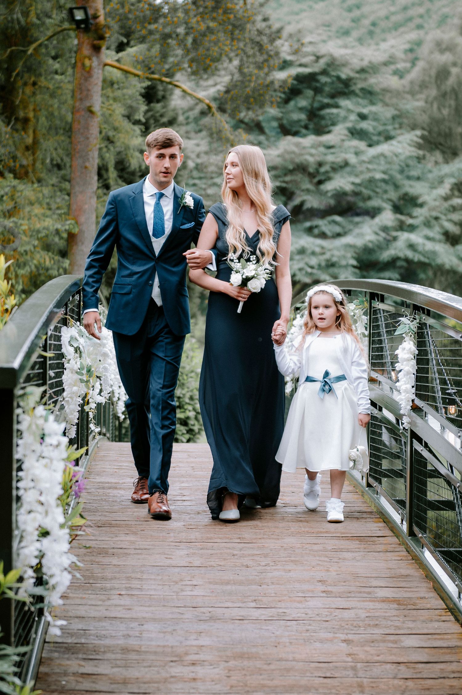 Wedding party members walking across a decorated wooden bridge in formal attire.