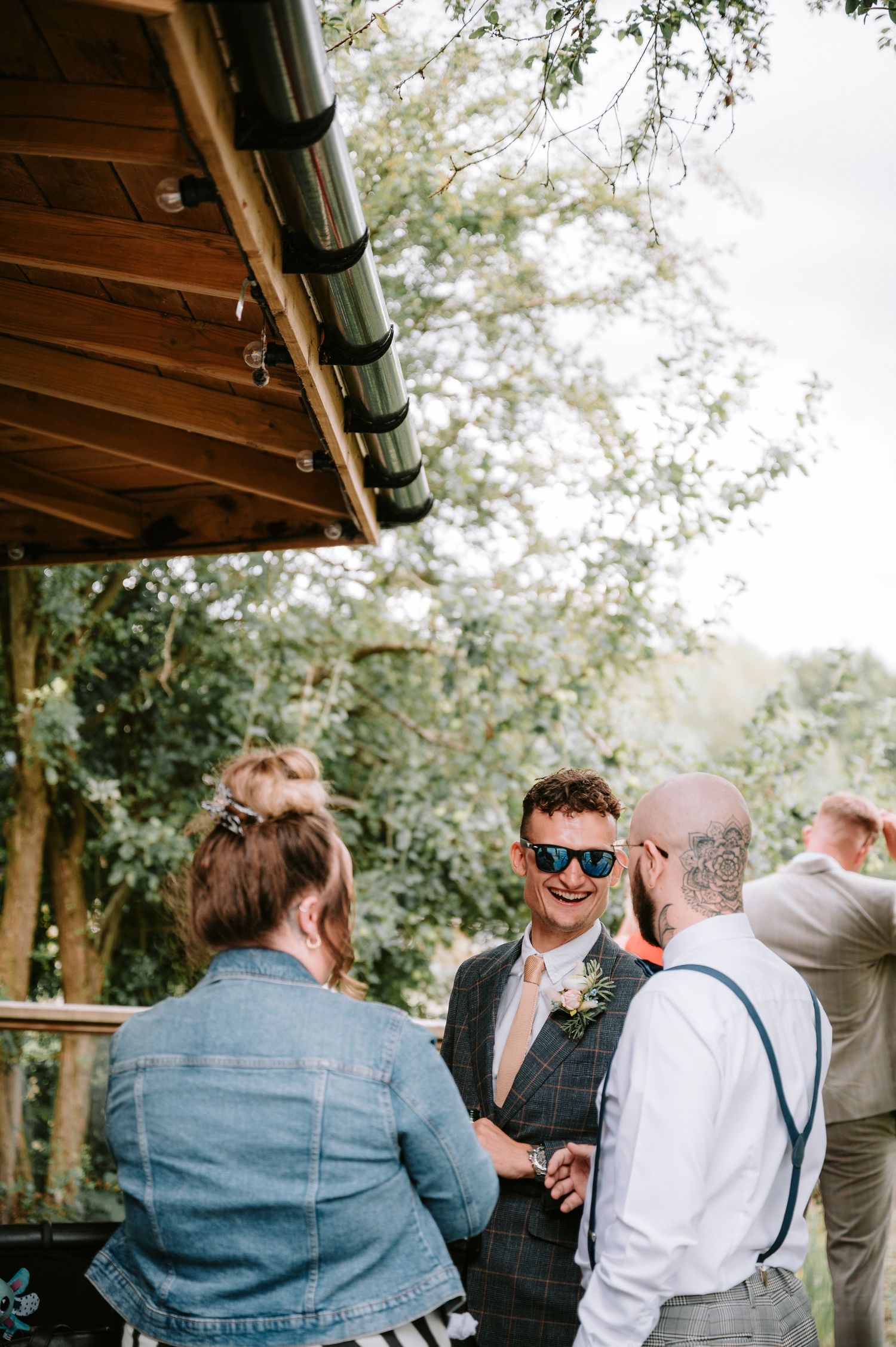Casual outdoor wedding gathering under wooden pavilion with guests in summer attire.