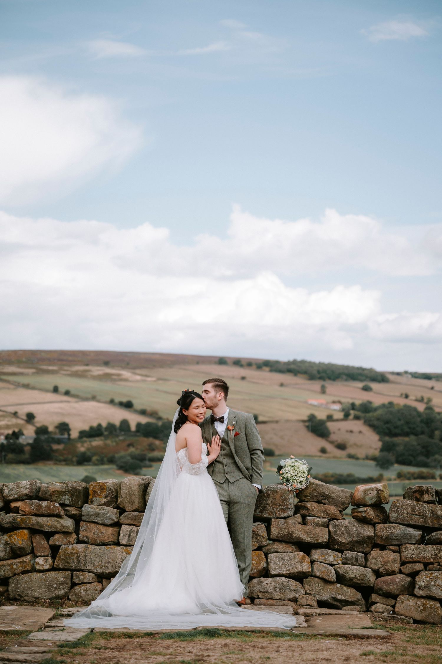 Wedding couple embraces against stone wall with rolling hills in background.