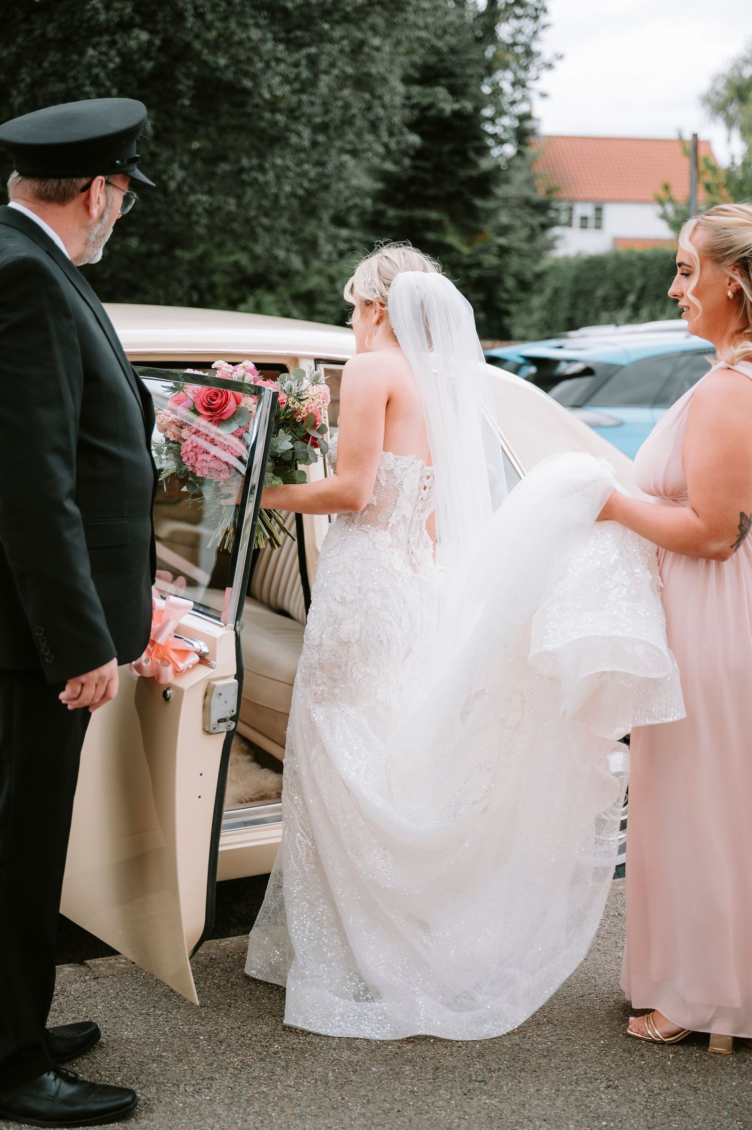 A bride in a white lace dress enters a classic car while being assisted by attendants on her wedding day.