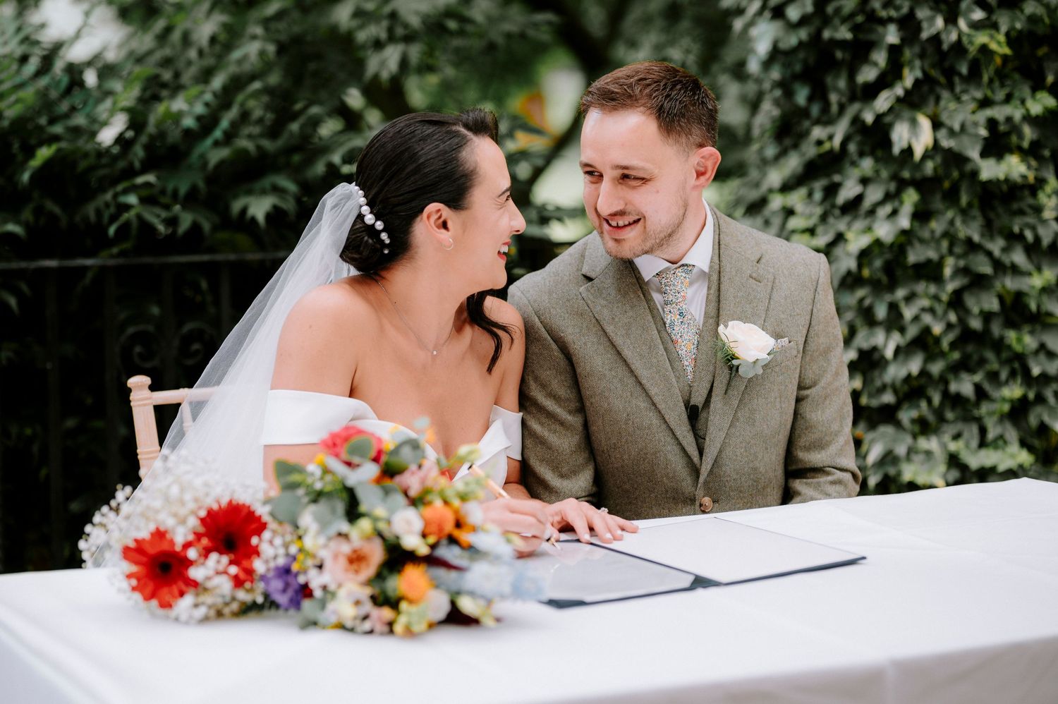Wedding couple signs marriage documents at outdoor ceremony with colorful floral bouquet on table.