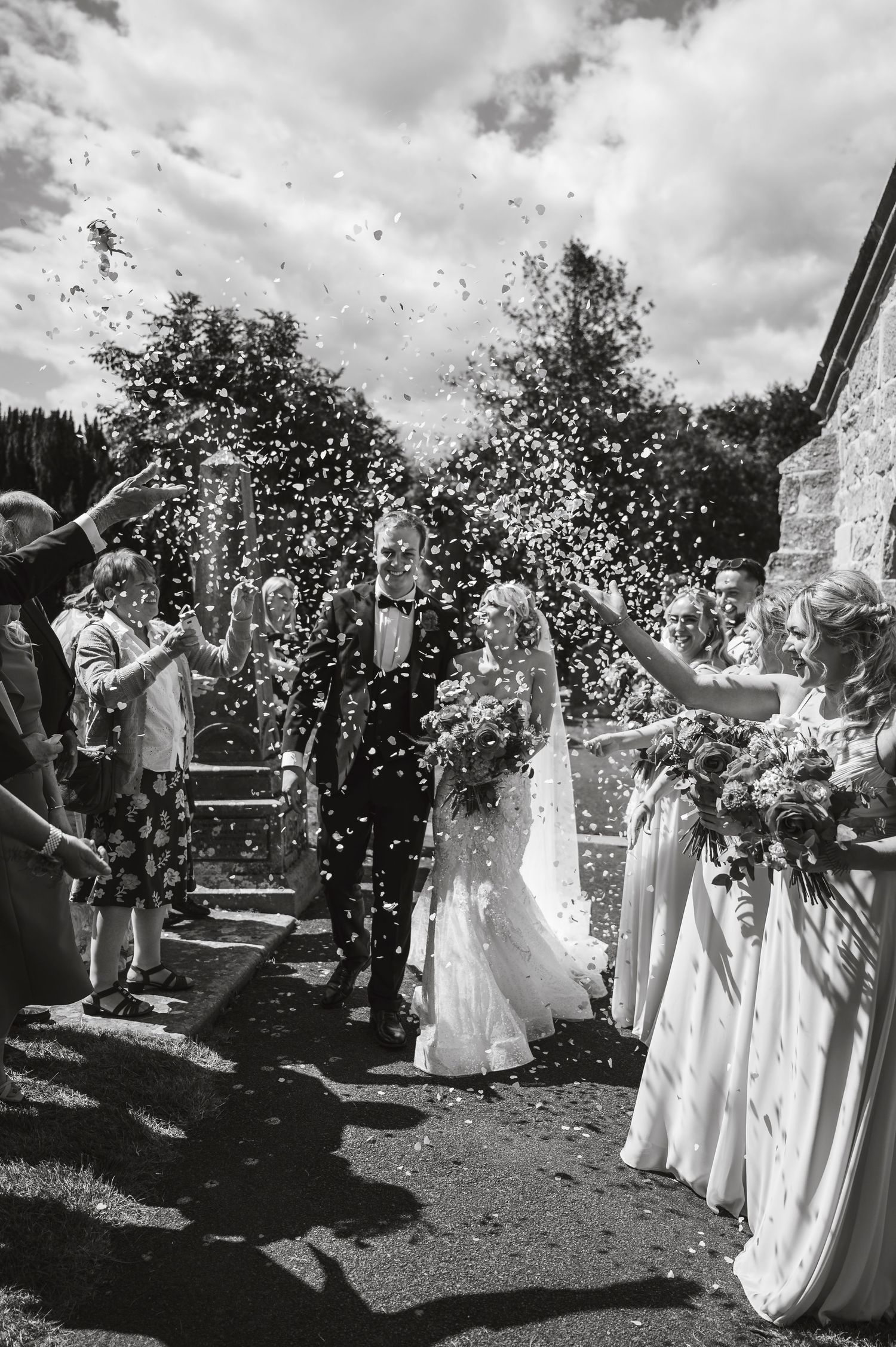 Black and white photo of wedding party celebrating with confetti outdoors.