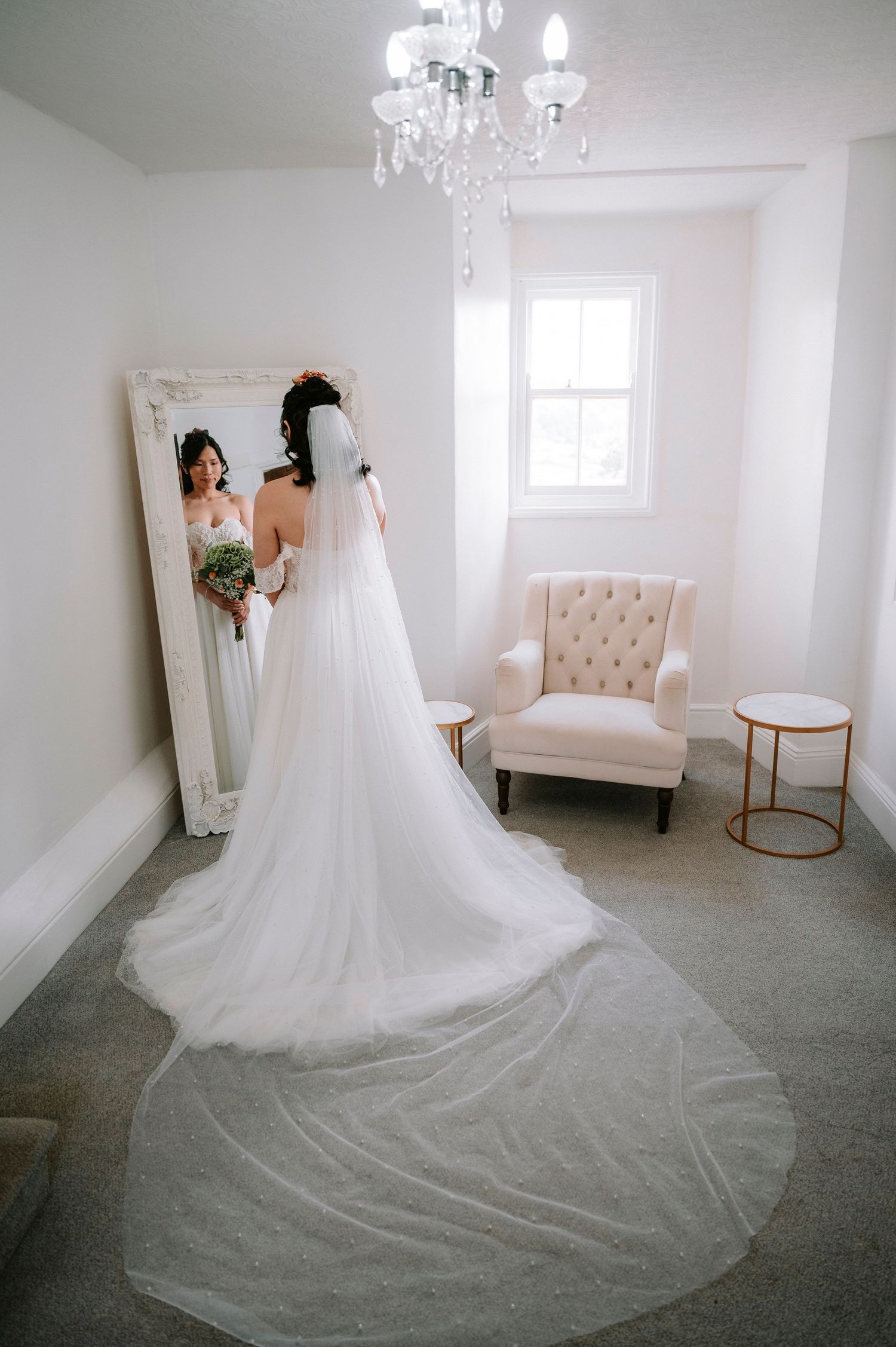 Flowing white wedding dress displayed in elegant bridal suite with chandelier and tufted furniture.