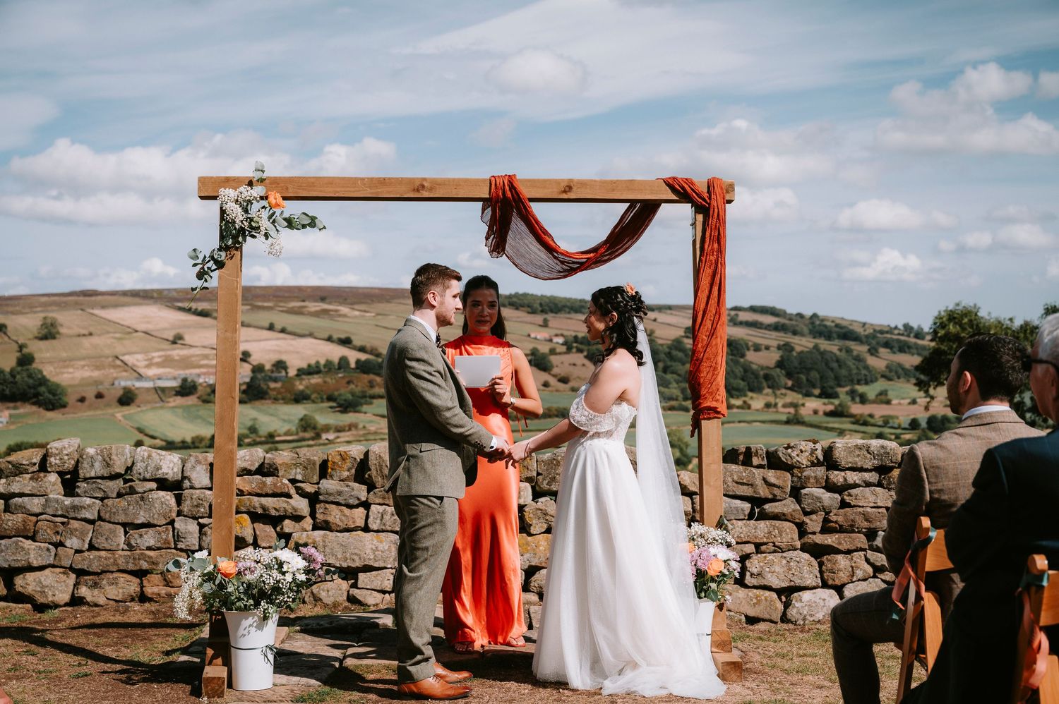 Outdoor wedding ceremony under a wooden arch with stone walls and rolling hills in the background.