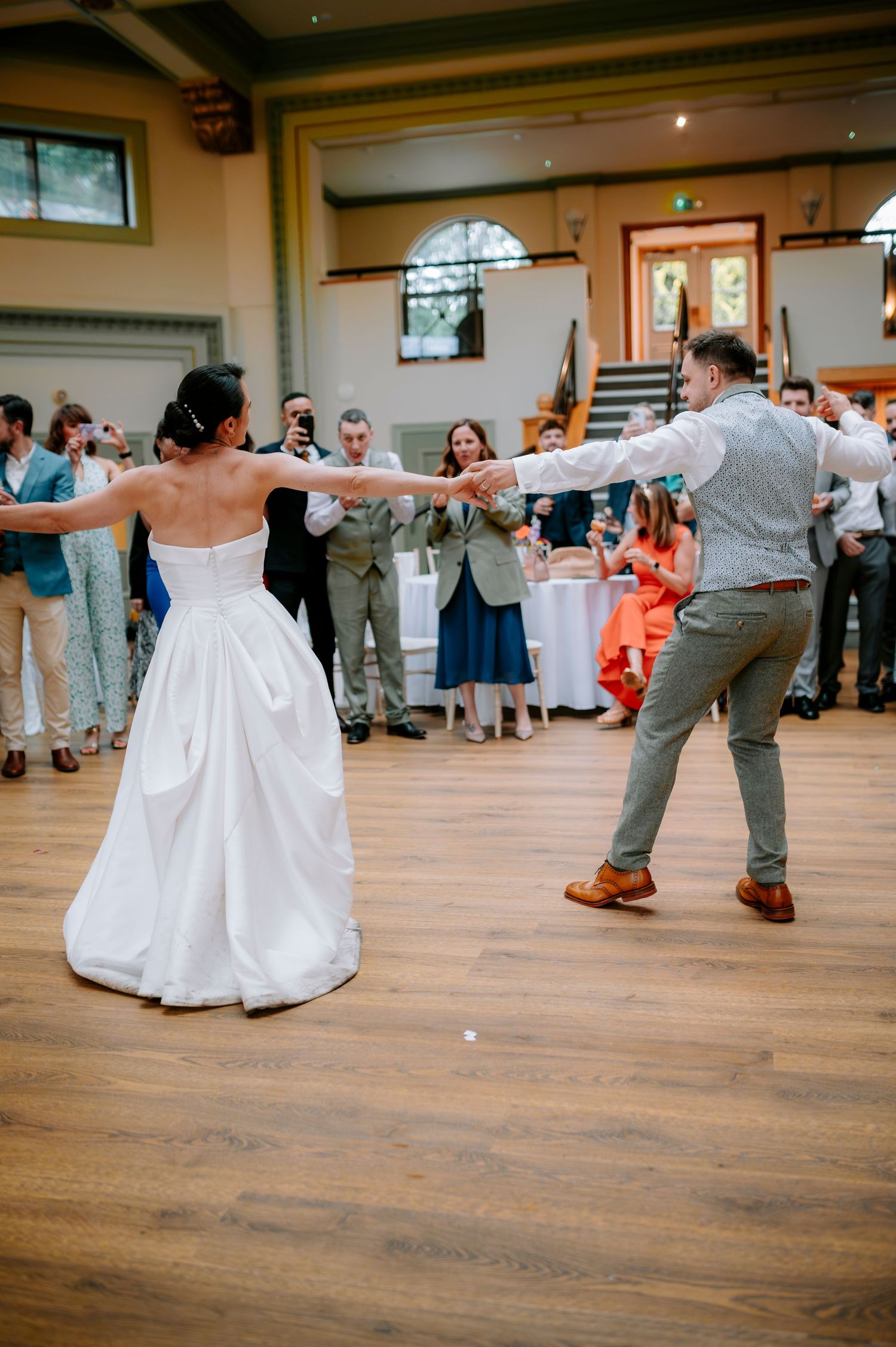 A couple performs their first dance in a reception hall with wooden floors and watching guests.