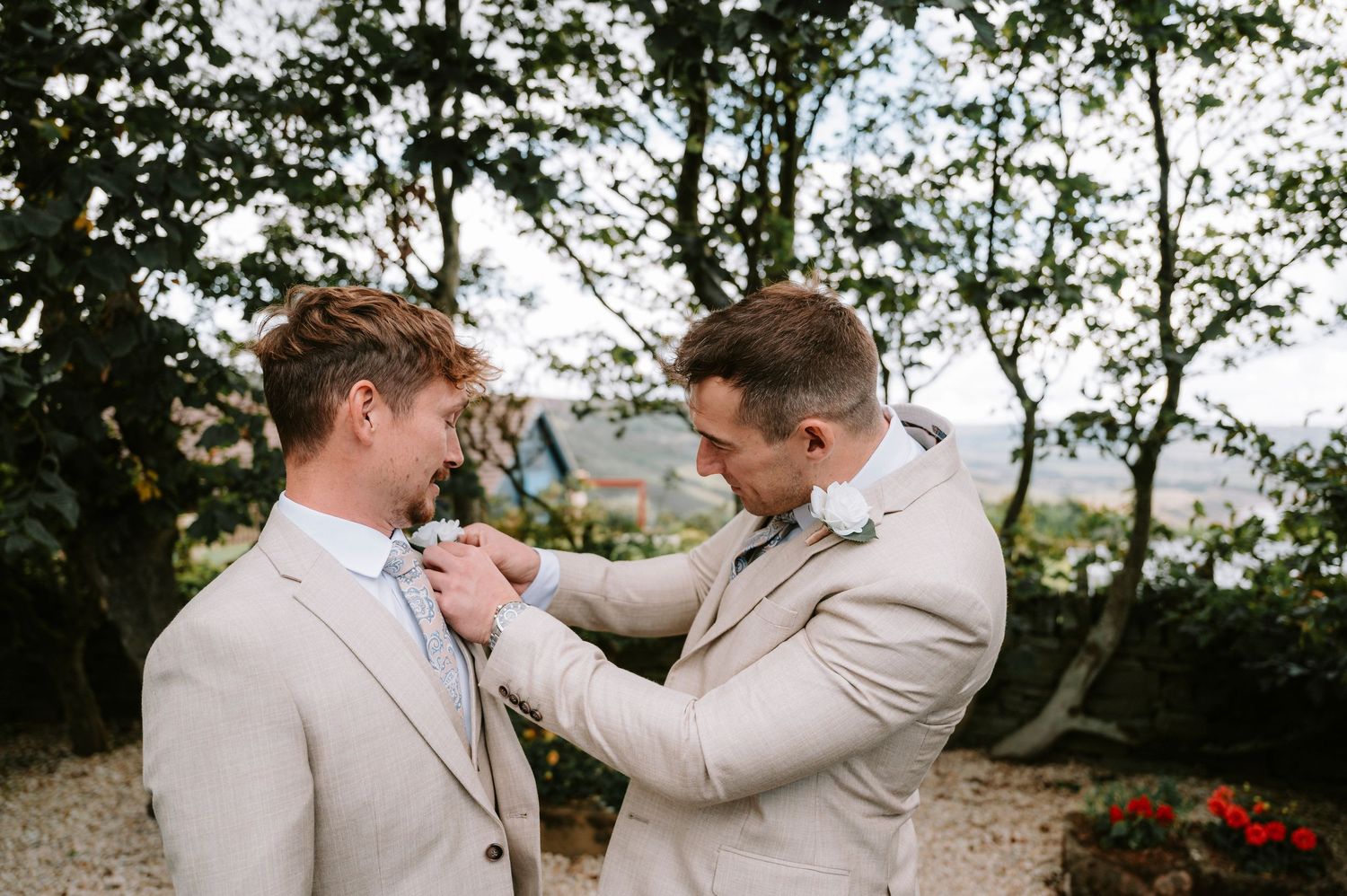 Two people in matching beige suits adjusting boutonniere outdoors.
