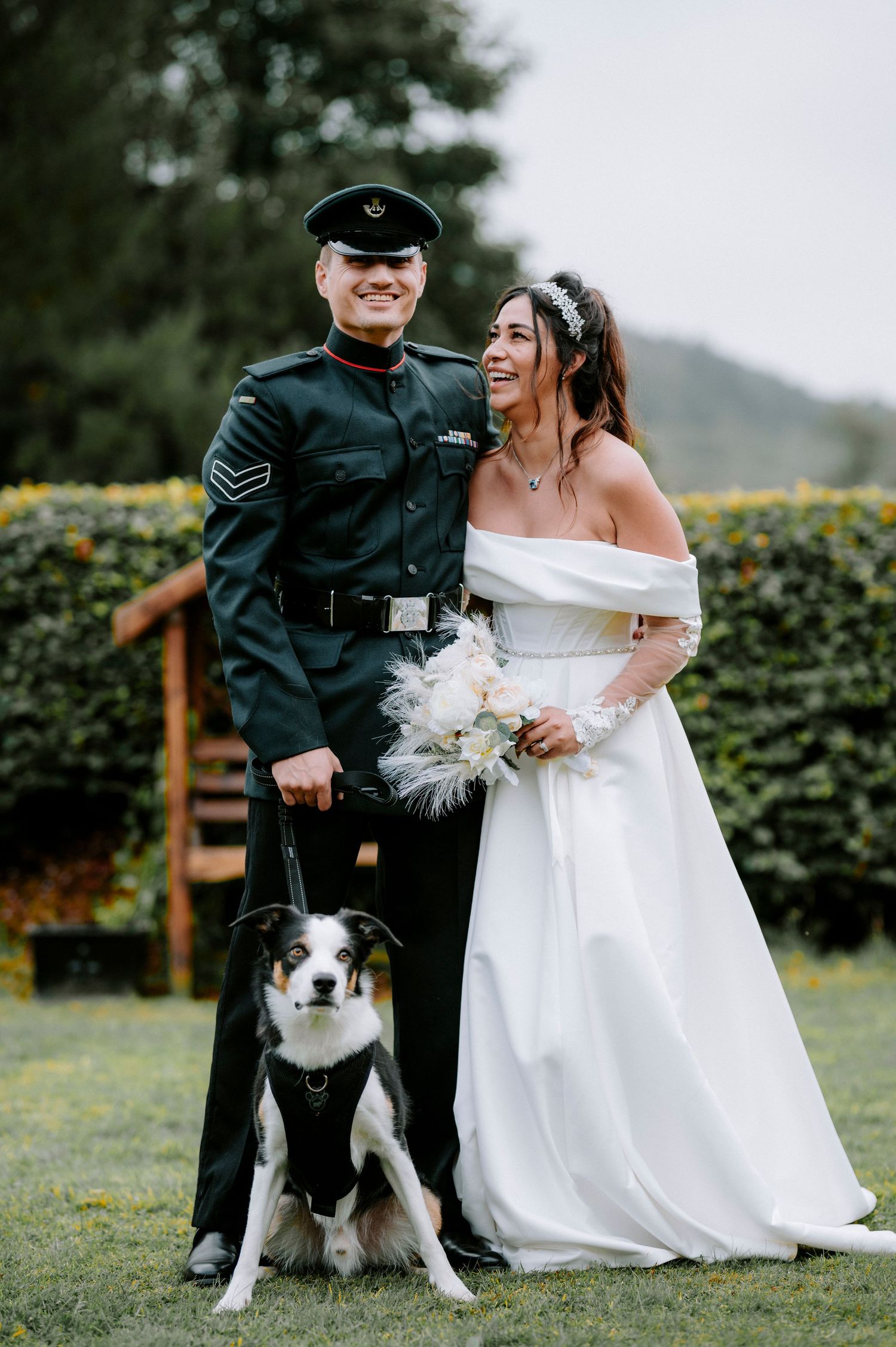 A wedding couple poses with their black and white border collie dog in an outdoor garden setting.