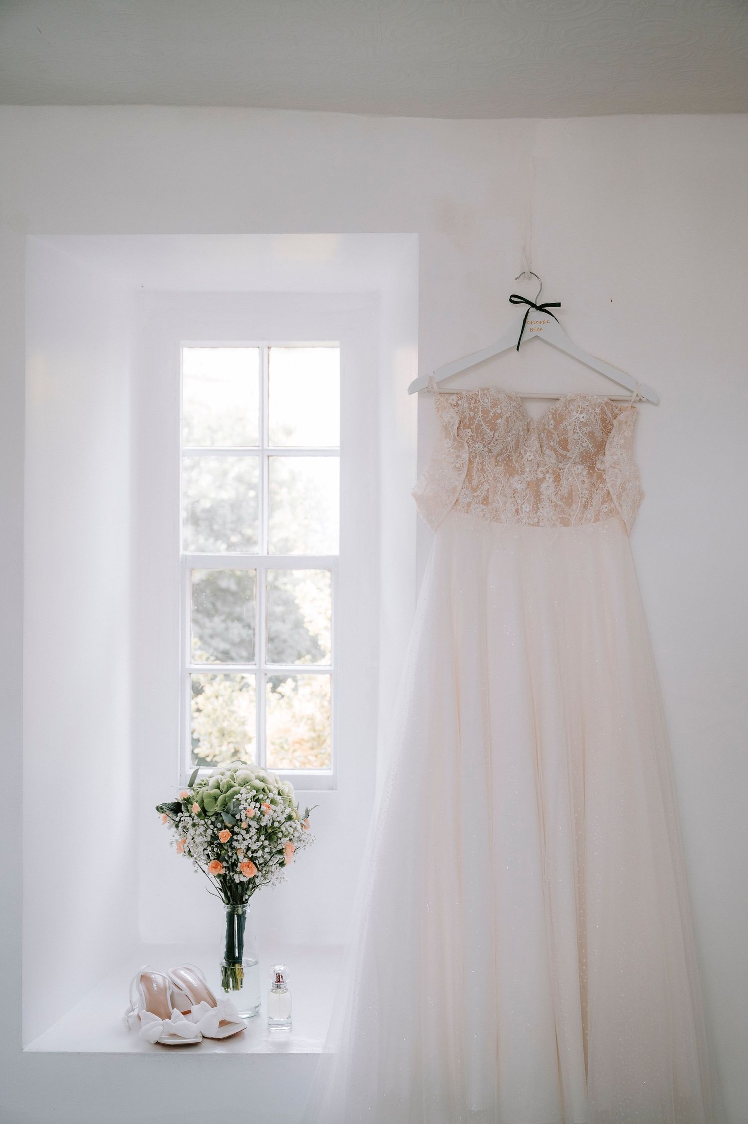 Elegant white wedding dress displayed near sunny window with floral arrangement.