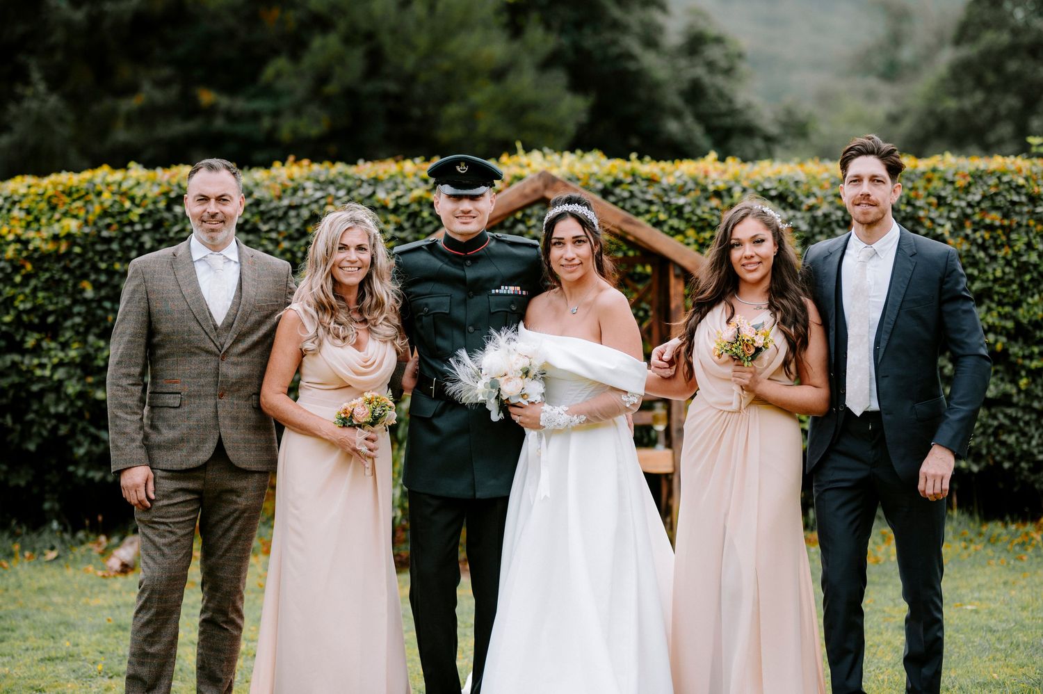 A wedding party in pastel attire poses outdoors against a backdrop of yellow flowers and greenery.