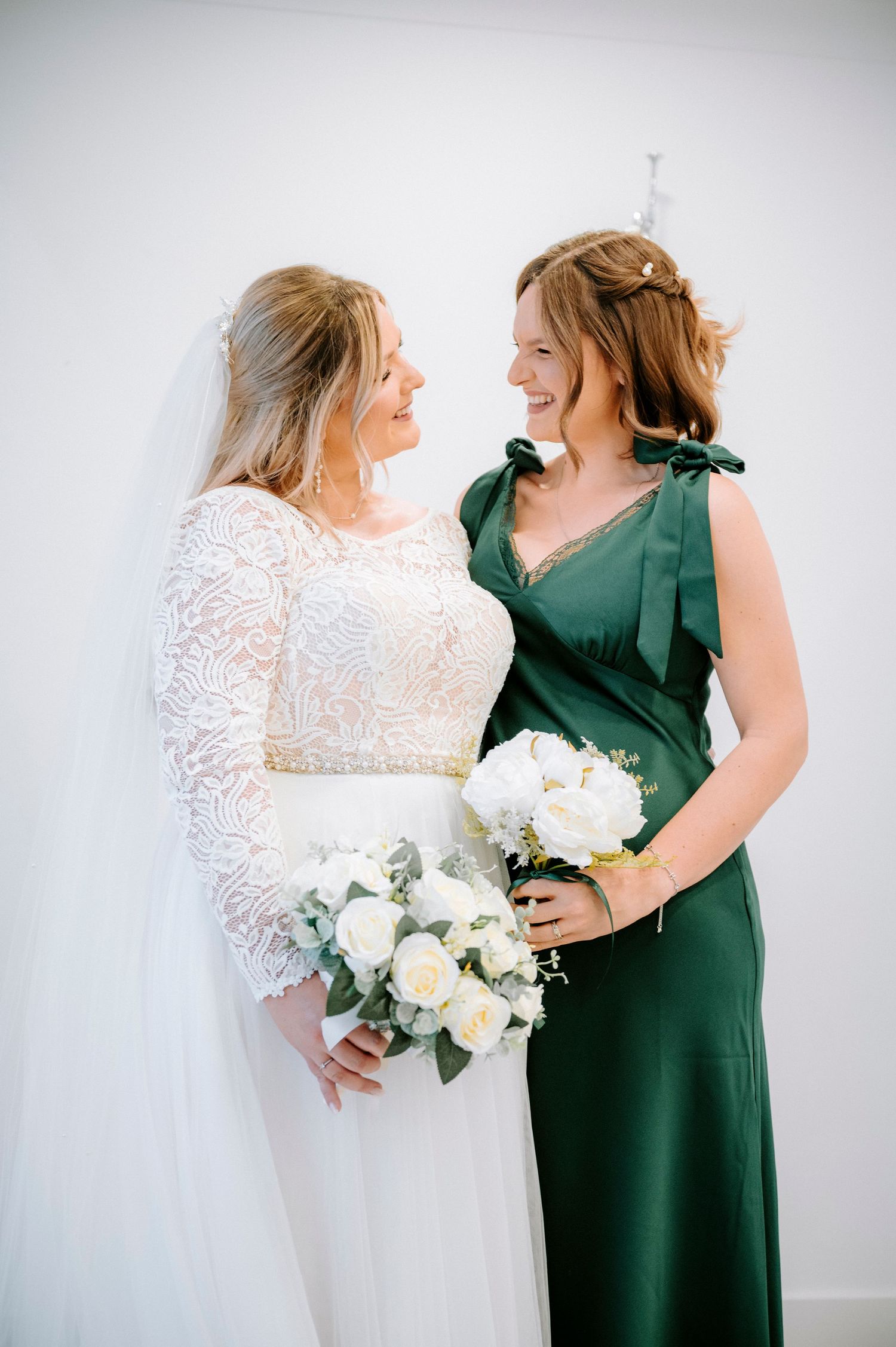 A bride in lace gown shares a joyful moment with bridesmaid wearing emerald green dress holding white bouquet.