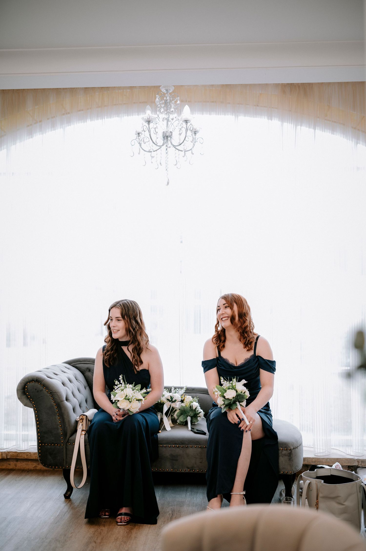 Bridesmaids in navy dresses sitting on grey couch with white bouquets.