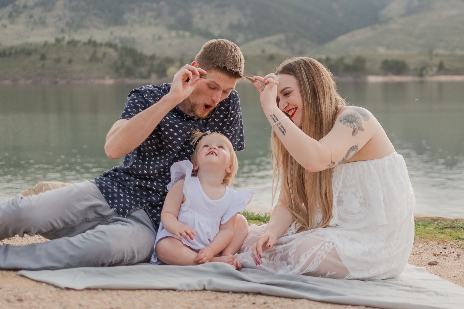 A young family sits on a blanket by a lake, parents playfully gesturing while their toddler looks up smiling. Fort Collin Family Photography