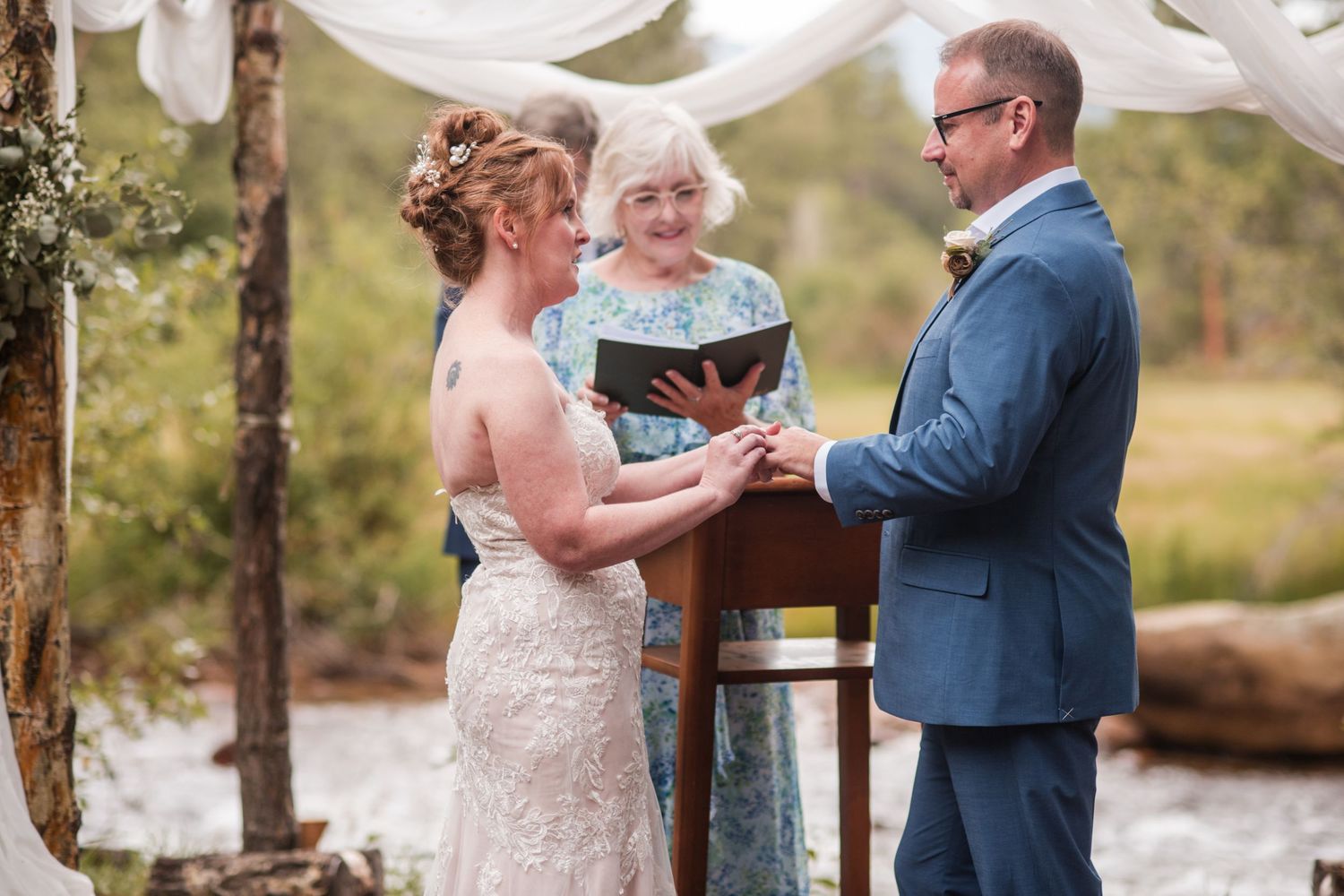 Bride and groom exchanging rings at an outdoor wedding ceremony with an officiant and guests in floral dresses. Riversong Inn Retreat
