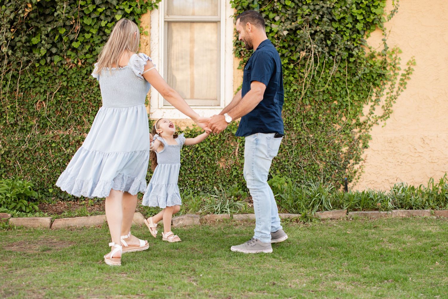 A happy family of three swinging their toddler daughter between them on a green lawn near an ivy-covered wall. Spring Family Photography