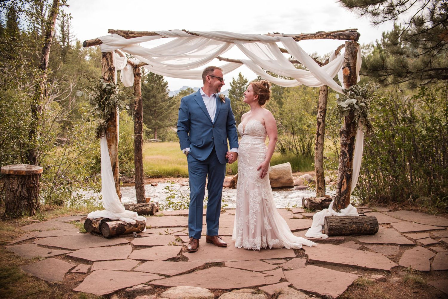 Bride and groom hold hands under rustic wooden arch with white draping at outdoor wedding ceremony in forest setting. Photography Angelice