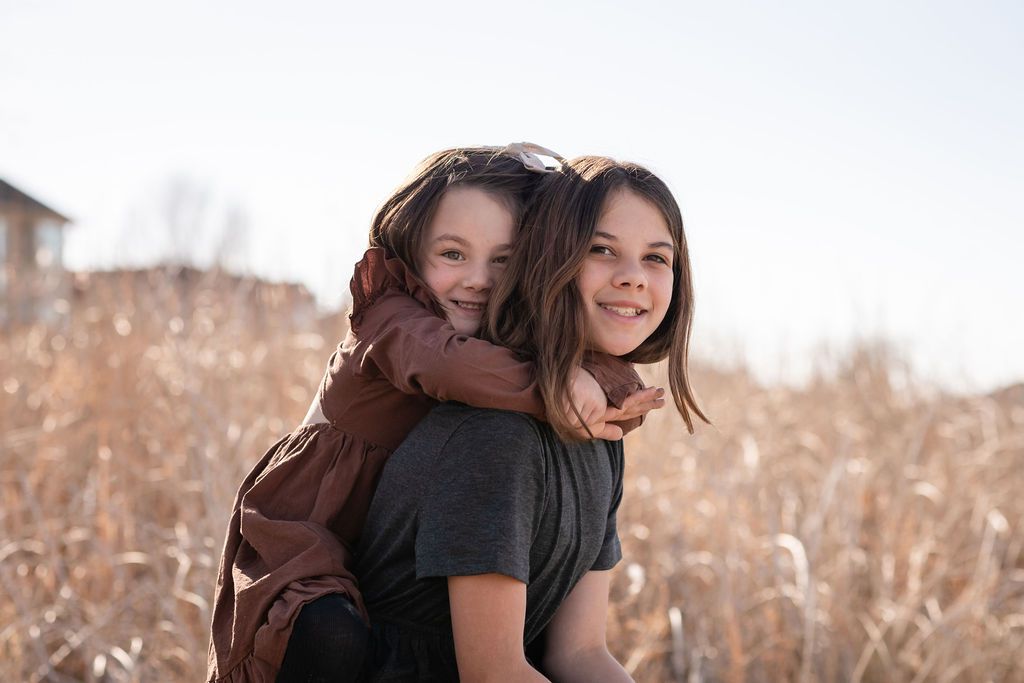 Two young girls smiling outdoors, one giving the other a piggyback ride, lakeside. Mcintosh Lake Longmont family photography