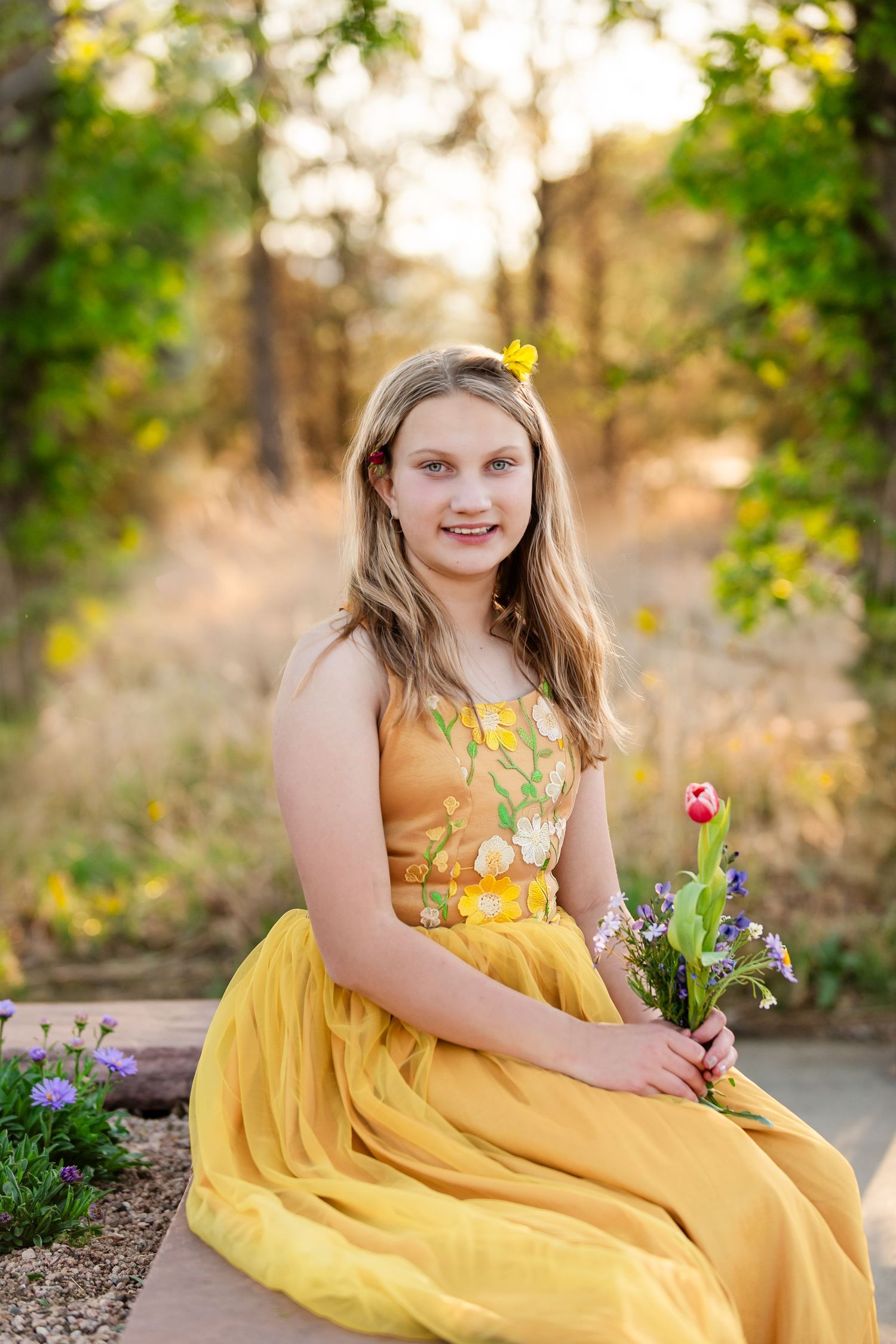 A young girl in a yellow floral dress sits outdoors holding a tulip bouquet, surrounded by greenery and wildflowers. Fine art photography