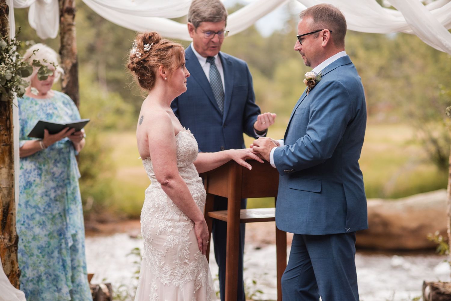 Bride and groom exchanging rings at outdoor wedding ceremony with officiant and guest in floral dress. Riversong Inn Retreat