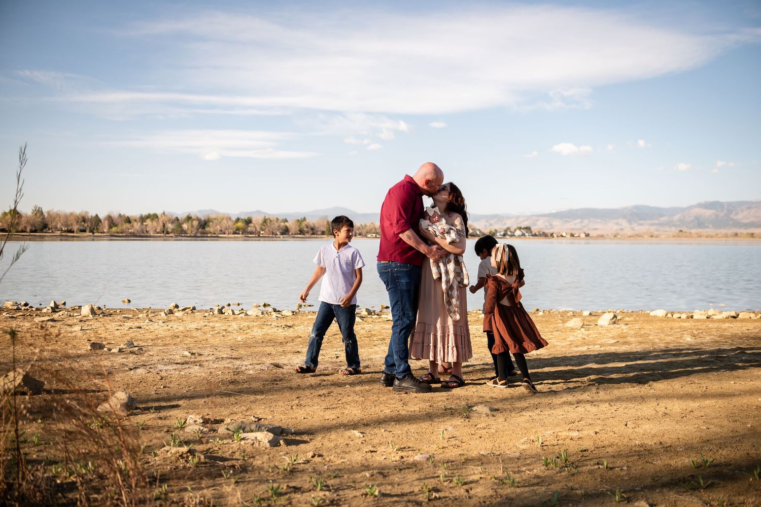 Family of four standing near a lake shore on a sunny day, parents embracing while children play nearby. Family Photography McIntosh Lake
