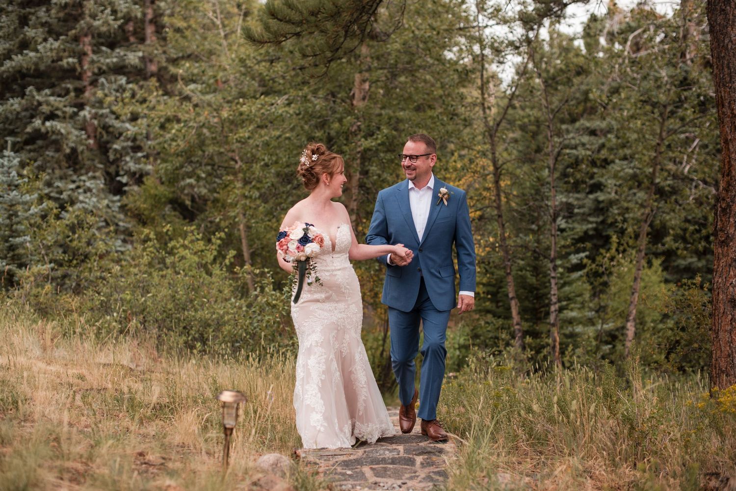 The bride in a lace gown and the groom in a blue suit walk hand in hand on a forest path surrounded by lush green trees, the first look