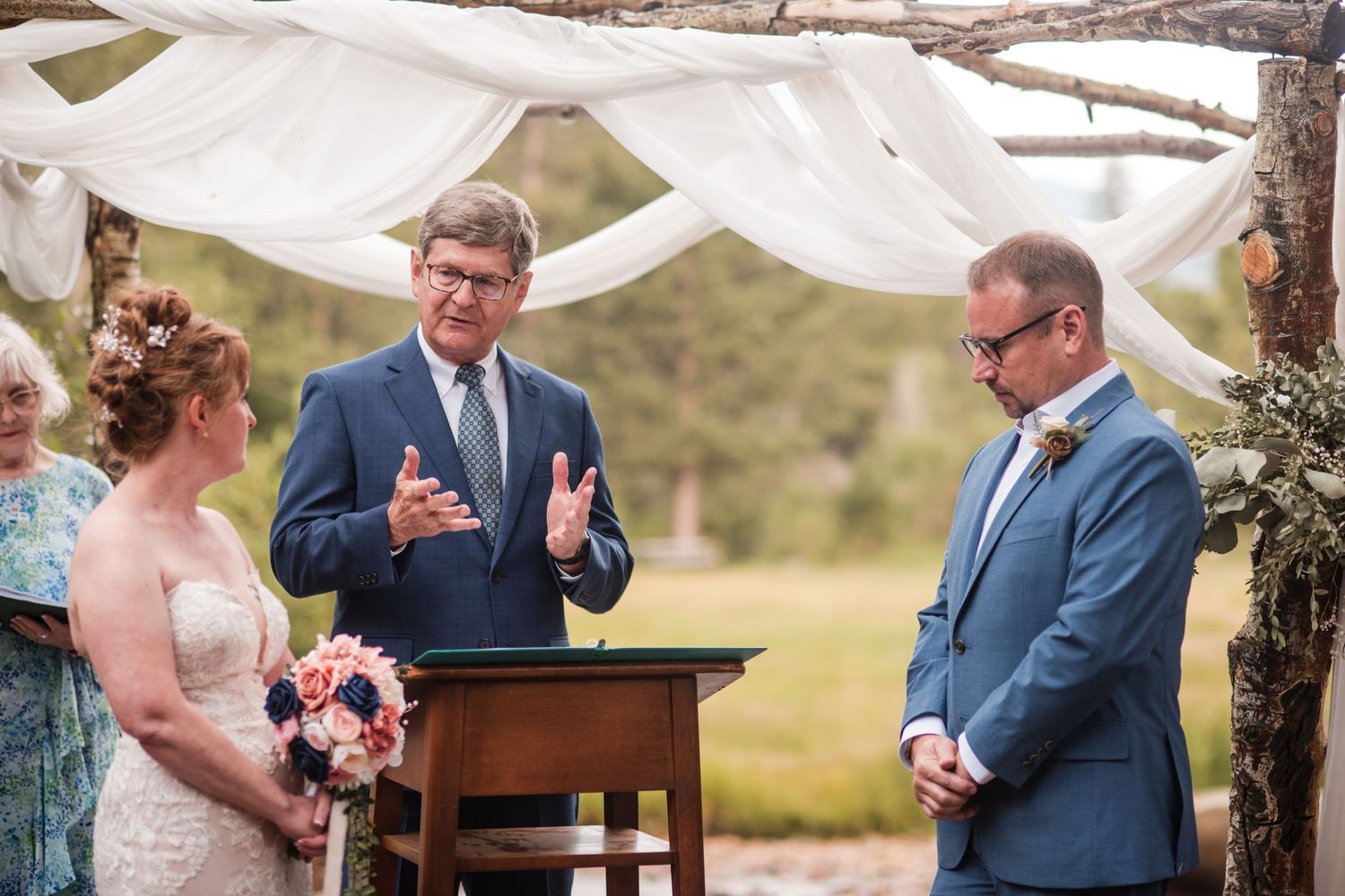 Outdoor wedding ceremony with bride, groom, and officiant under a rustic wooden arch draped with white fabric.