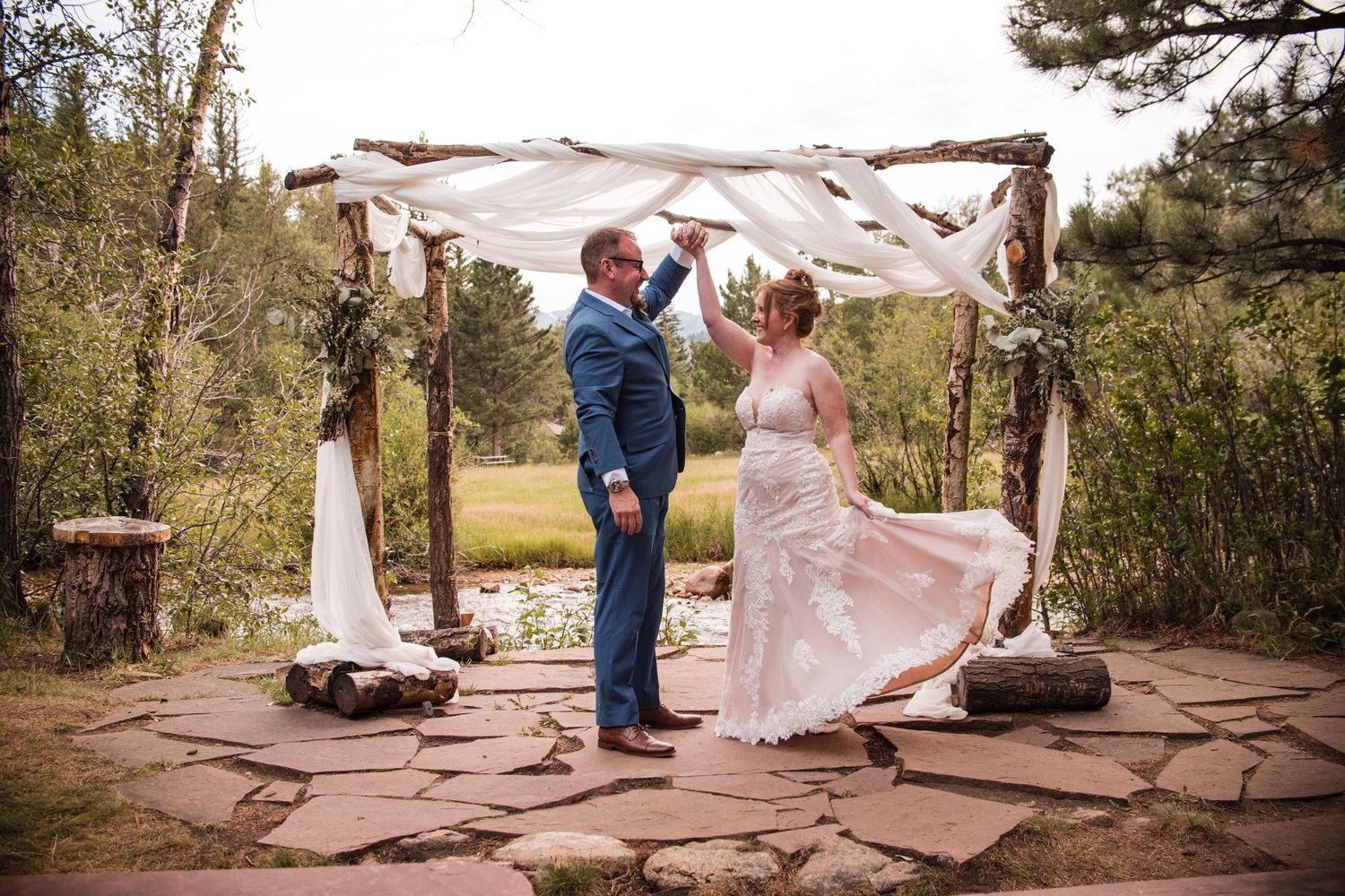 Bride and groom dancing under rustic wooden arch with white draping at outdoor wedding ceremony in forest setting.RiversongInnRetreat-Estes